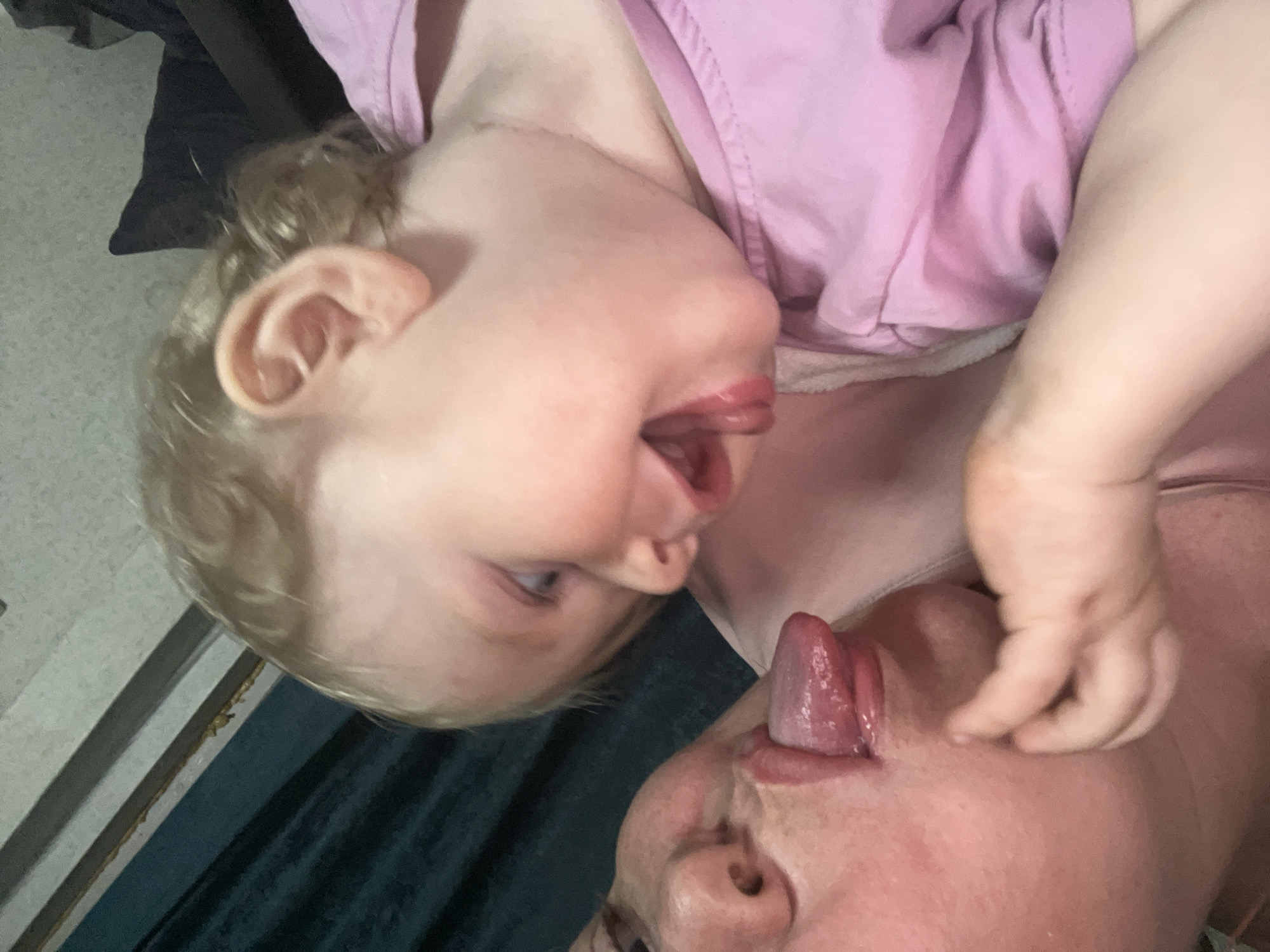 The image shows a close-up of two people, likely an adult and a child, interacting. The child, with blonde hair, is wearing a pink shirt and is positioned above the adult. The child's face is turned downwards, with their mouth open and tongue sticking out. The adult's face is visible from below, also with their tongue extended towards the child's. The scene is intimate and playful, suggesting a moment of bonding or silliness between the two individuals. The lighting is soft, and there are no prominent background details that would indicate a specific time of day or weather conditions. The setting appears to be indoors, possibly a home environment, given the casual nature of the interaction and clothing. There is no visible text in the image. The emotion conveyed is one of joy and affection.