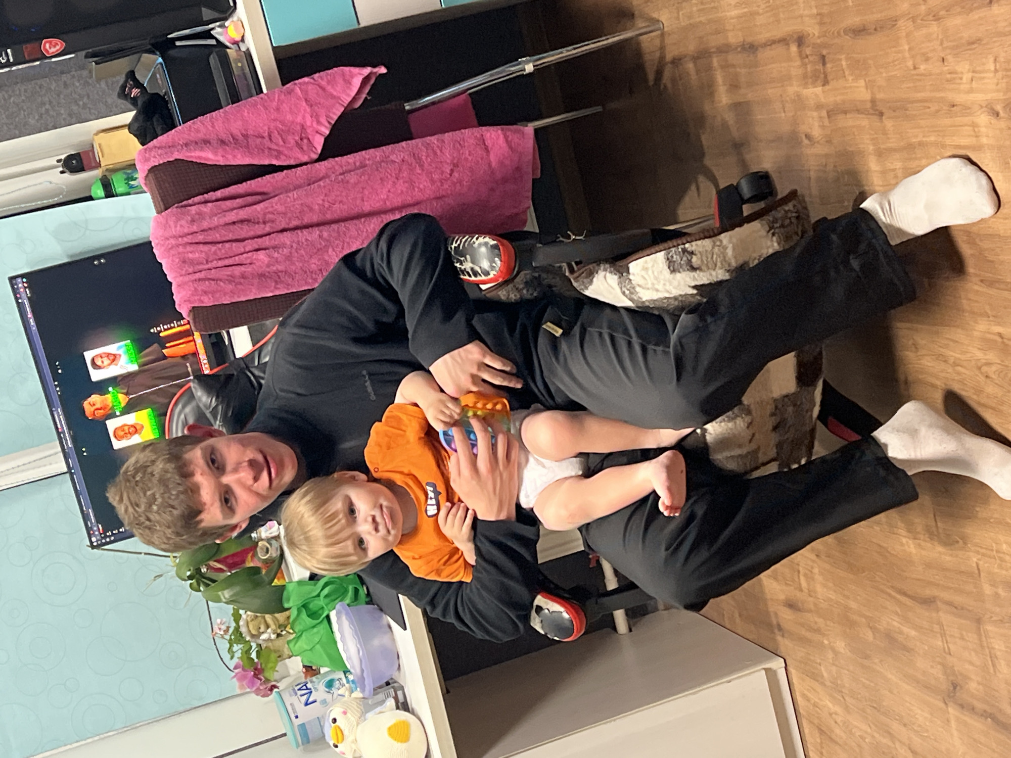 In Noviy, Russia, a young man with short brown hair sits in a gaming chair, holding a toddler on his lap. The toddler, with blonde hair, wears an orange t-shirt and a diaper, and is playing with a colorful bubble toy. The man is dressed in a black turtleneck and black pants, and both are wearing white socks. The background shows a desk with a computer monitor displaying a video call with multiple participants, some of whom appear to be rendered as caricatures. Various items are visible on the desk, including a pink towel draped over the chair, a plant with pink flowers, and several toy animals. The room has wooden flooring.
