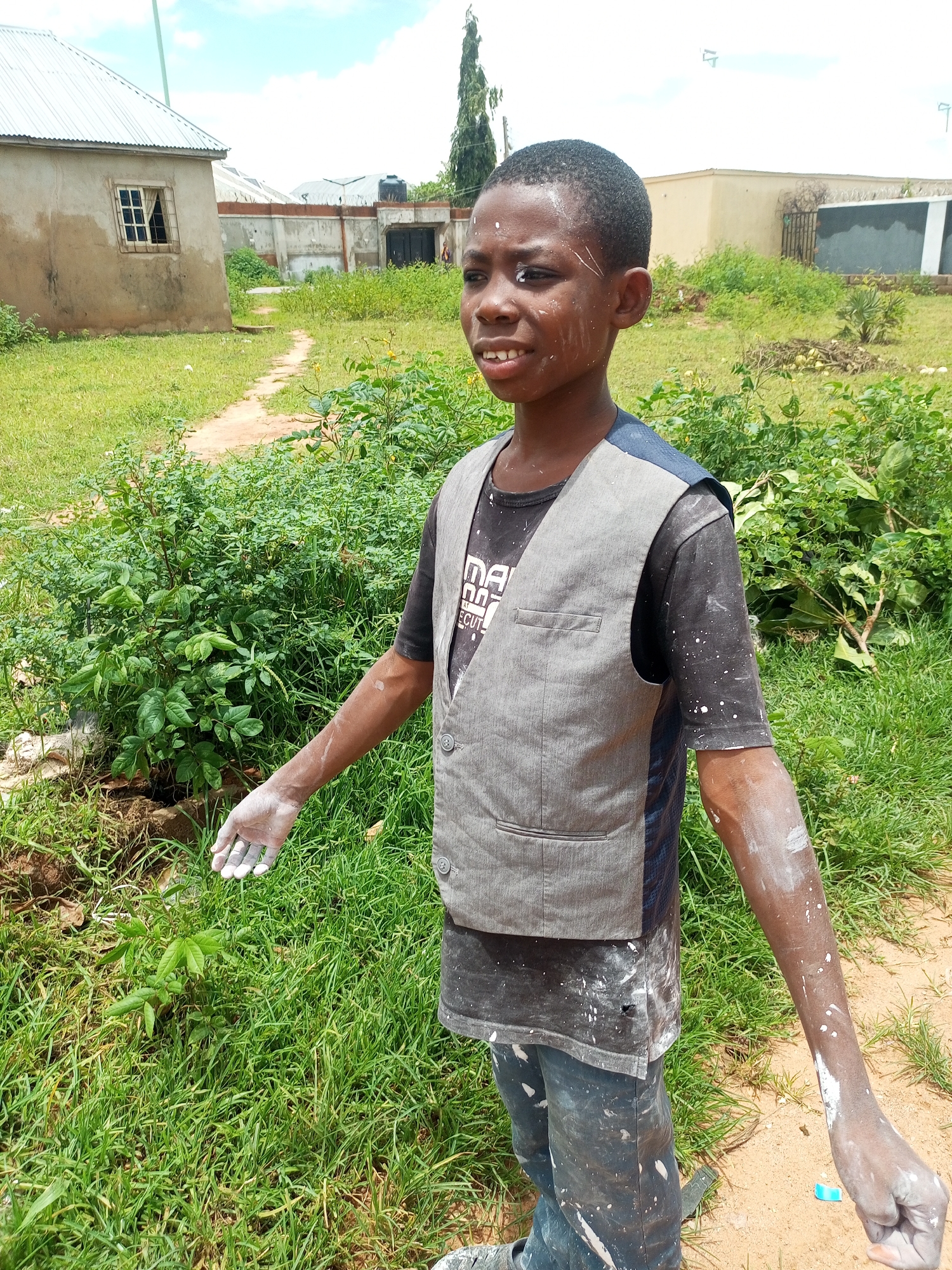 A young Black male stands in a grassy outdoor area, his skin and clothing smudged with white paint. He is wearing a dark t-shirt with a grey vest over it and dark pants. His left arm is extended, and his hand is open and covered in white paint. The background shows several buildings and lush green vegetation, typical of a suburban or semi-rural area. The sky is partly cloudy. The scene suggests he may have been engaged in painting or a similar activity. This image was taken in Tempure, Nigeria.
