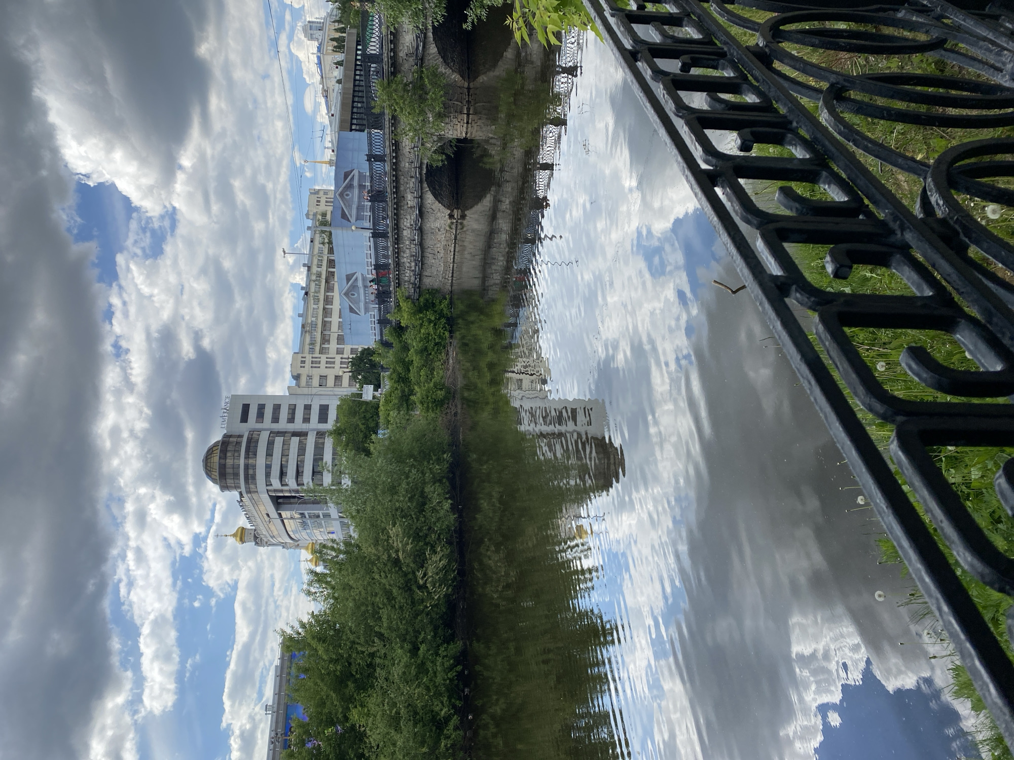 The image depicts a scene in Yekaterinburg, Russia, specifically the Iset River and the surrounding architecture. In the foreground, a black wrought iron fence runs along the edge of the riverbank. Beyond the fence, the Iset River reflects the sky and clouds, creating a mirrored effect. On the opposite bank, a line of trees borders the river, and behind the trees, various buildings are visible, including modern office structures and older buildings with distinctive architectural features. A bridge with arched supports spans the river. The sky above is filled with white clouds.
