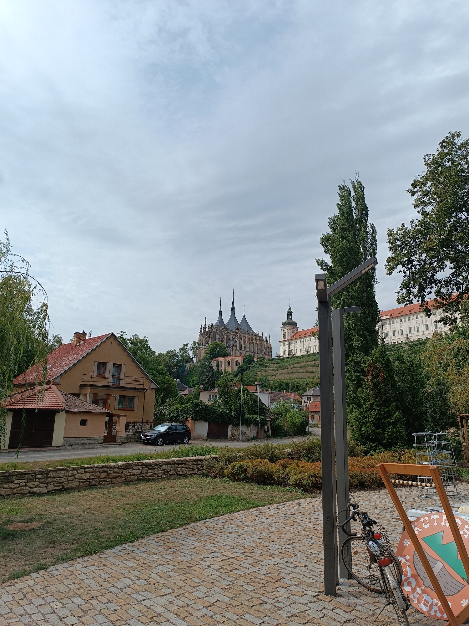 The image shows a street scene in Kutná Hora, Czechia, with the iconic Church of Saint Barbara in the background. In the foreground, a paved walkway made of light brown and gray cobblestones leads into the scene. To the left, a low stone wall separates the walkway from a grassy area with sparse dry grass and some bushes. Beyond the grassy area, a residential building with an orange-tiled roof and a garage is visible. A dark-colored car is parked on the street in front of the house.
Further back, the magnificent Gothic Cathedral of Saint Barbara dominates the hilltop, its spires reaching towards a cloudy sky. Adjacent to the cathedral, on a terraced slope, is a large, white building with red-tiled roofs and numerous windows, likely part of the former Jesuit college. Vineyards are visible on the slopes leading up to the buildings.
On the right side of the foreground, a tall, slender modern street lamp stands next to a bicycle leaning against it. A wooden frame of an outdoor game, possibly a quoits or ring toss game, is also present, decorated with a circular graphic featuring wings and ornate patterns. Lush green trees frame the right side of the scene, with one particularly tall, thin tree reaching high into the sky. The overall atmosphere is serene and captures a picturesque view of the historic city.