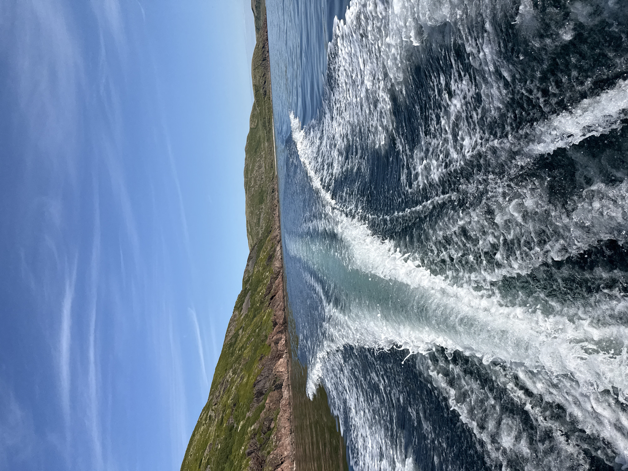 The image shows a view from the stern of a moving boat, looking back at the wake. The water is a deep blue, with white foam and turbulence forming a prominent V-shape behind the vessel. In the distance, a rugged coastline with green, rocky hills rises from the water. The sky is a clear blue with a few wispy white clouds. The scene is likely located on the coast of Teriberka, Russia, a settlement situated on the Kola Peninsula along the Barents Sea, known for its stark natural beauty. There are no people or specific buildings visible in this shot, focusing solely on the natural environment and the effect of the boat's passage.
