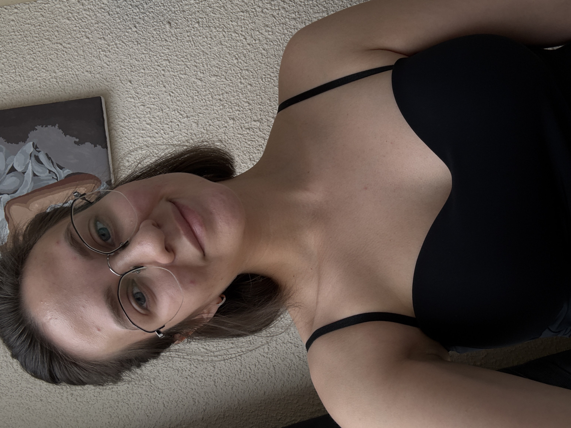 A young woman with brown hair and glasses is posing for a selfie indoors, against a textured wall. She is wearing a black strapless top. A framed artwork hangs on the wall behind her. The lighting suggests an indoor setting, possibly a room with natural light.