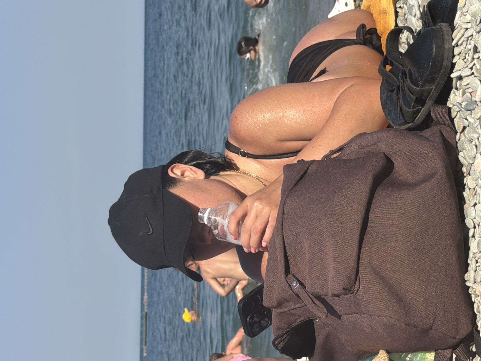 A woman wearing a black baseball cap and a black bikini lies on a pebble beach with her back to the camera. She is holding a clear plastic bottle of drink up to her mouth. A brown backpack is in the foreground to her left, and a pair of black sandals are to her right. The sea is behind her, with several people swimming in the water. The sun is shining brightly, creating a warm atmosphere. The location is the city of Partenit, Ukraine.