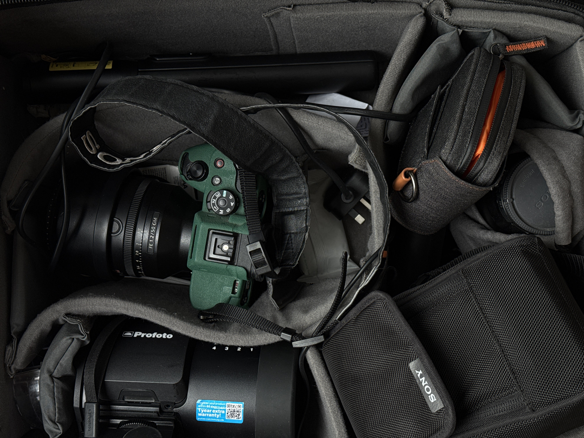 This media file presents an overhead view of various professional photography equipment neatly organized within a dark gray camera bag, suggestive of preparation for a shoot or travel.
The central subject is a camera body, possibly a Sony Alpha series, encased in a distinctive green protective silicone skin. A large lens, identified by the visible text "FE 1.2/50 GM", is attached to the camera. The camera's mode dial shows settings such as "M", "A", "S", "AUTO", "C1", "C2", and "MENU". A dark, worn camera strap with partially visible lettering (likely "SONY") is draped over the camera.
Below the camera and lens, a large black cylindrical Profoto lighting component is visible, bearing the "Profoto" logo and a sticker that reads "Register your product within 60 days to get 1 year extra warranty!" and "profoto.com/setupguide", along with numerical markings "4 3 2 1". To the right, a small, dark gray zippered pouch with an orange zipper and accents is nestled among the bag's dividers, likely holding accessories. Next to this pouch, a black Sony E-mount lens cap is visible. Another black fabric pouch or case, labeled "SONY", occupies the bottom right corner of the bag. Various cables and other minor accessories are tucked between the padded compartments.
The scene conveys a sense of preparedness and organization for a photography assignment, with essential gear securely packed. While the location context is Dubai, UAE, no specific visual cues in the image indicate this geographical setting.