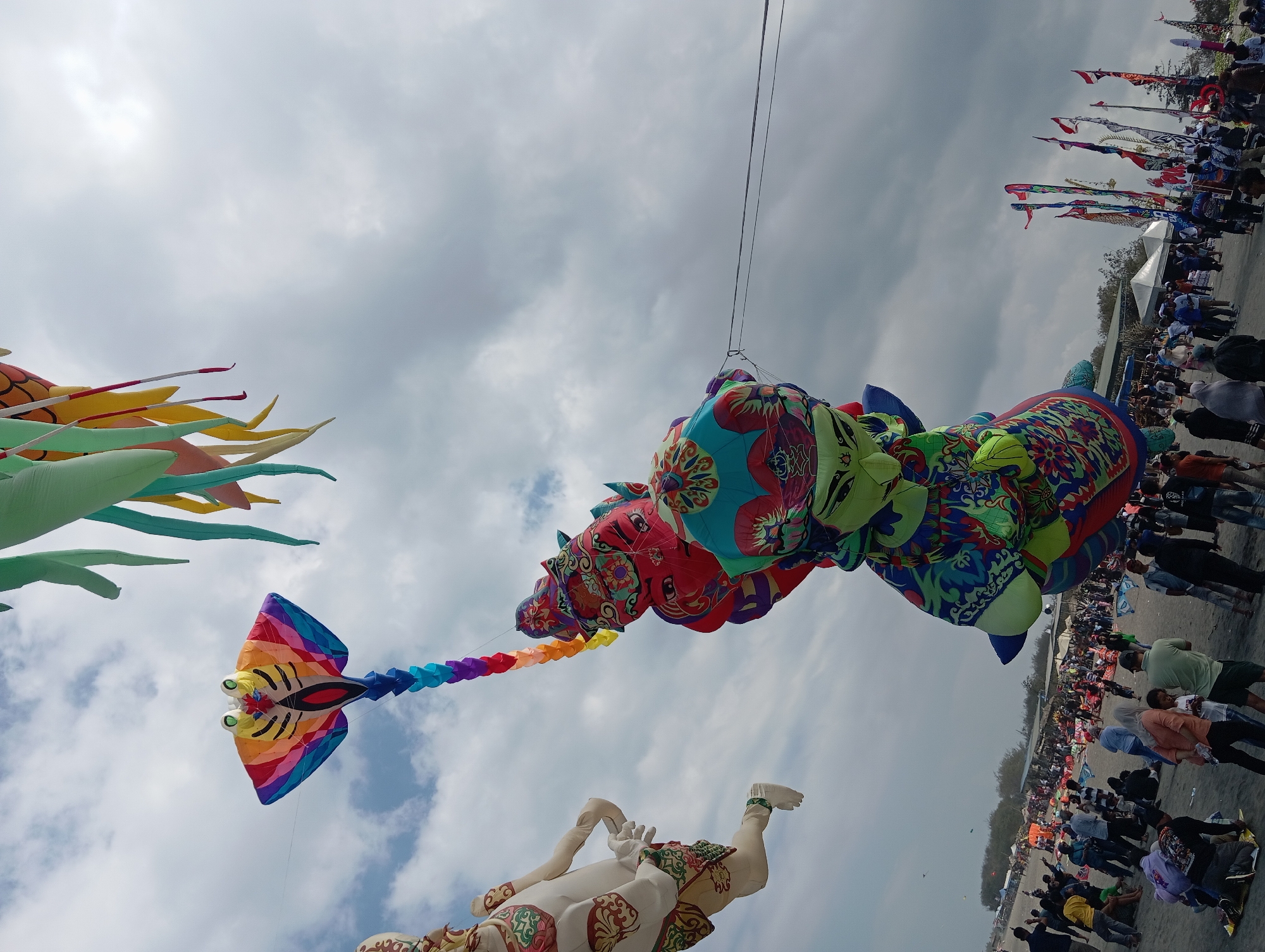 This is a low-angle, medium shot of a kite festival in Parangtritis, Indonesia, under a partly cloudy sky. The foreground is dominated by a very large, colorful kite resembling a stingray with a rainbow tail, and another kite shaped like a humanoid figure adorned with intricate patterns. Behind these kites, a crowd of people mills about on a sandy beach, interspersed with stalls and flags. The atmosphere appears festive, with many individuals observing the kites. The lighting is diffused, suggesting an overcast day.