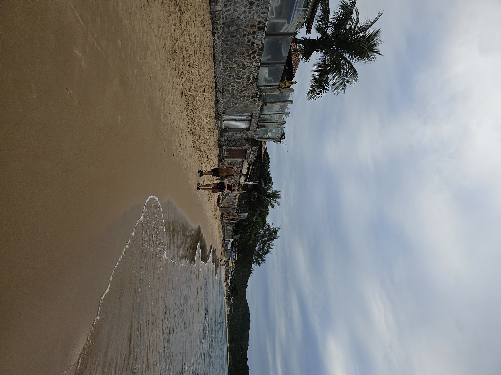 The image shows a coastal scene in Armação Dos Búzios, Brazil. A sandy beach stretches along the left side of the frame, meeting the ocean's edge where waves are visible. Adjacent to the beach is a stone structure with windows, likely a building or part of a coastal property. A palm tree extends into the sky, which is partially covered with clouds. Two people are walking on the beach near the stone structure. Further along the coast, there is vegetation, including trees and other greenery. The sky shows a mix of white and gray clouds.