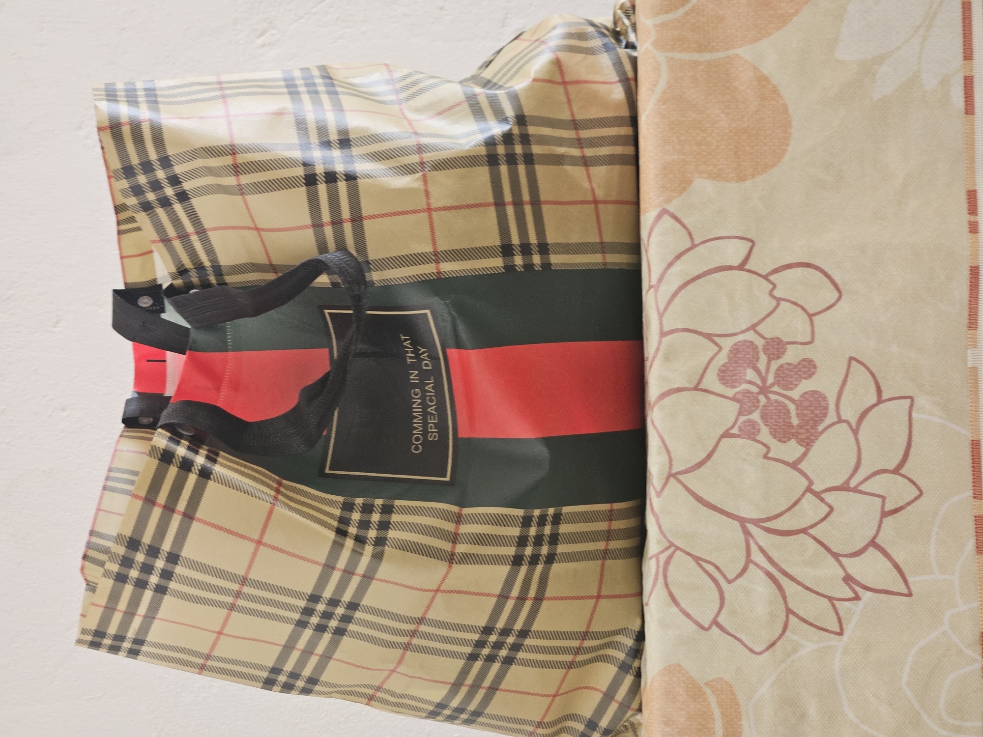 A tan, plaid tote bag with black straps rests on a surface covered with a patterned fabric. The tote bag has a green panel with two vertical red stripes and a black rectangular label in the center. The label has white text that reads "COMMING IN THAT SPEACIAL DAY". The fabric underneath the bag features a beige background with large, stylized floral patterns in pink and orange. The scene is illuminated by soft lighting, casting subtle shadows. The background wall appears to be textured and white. This image is located in Saray, Azerbaijan.