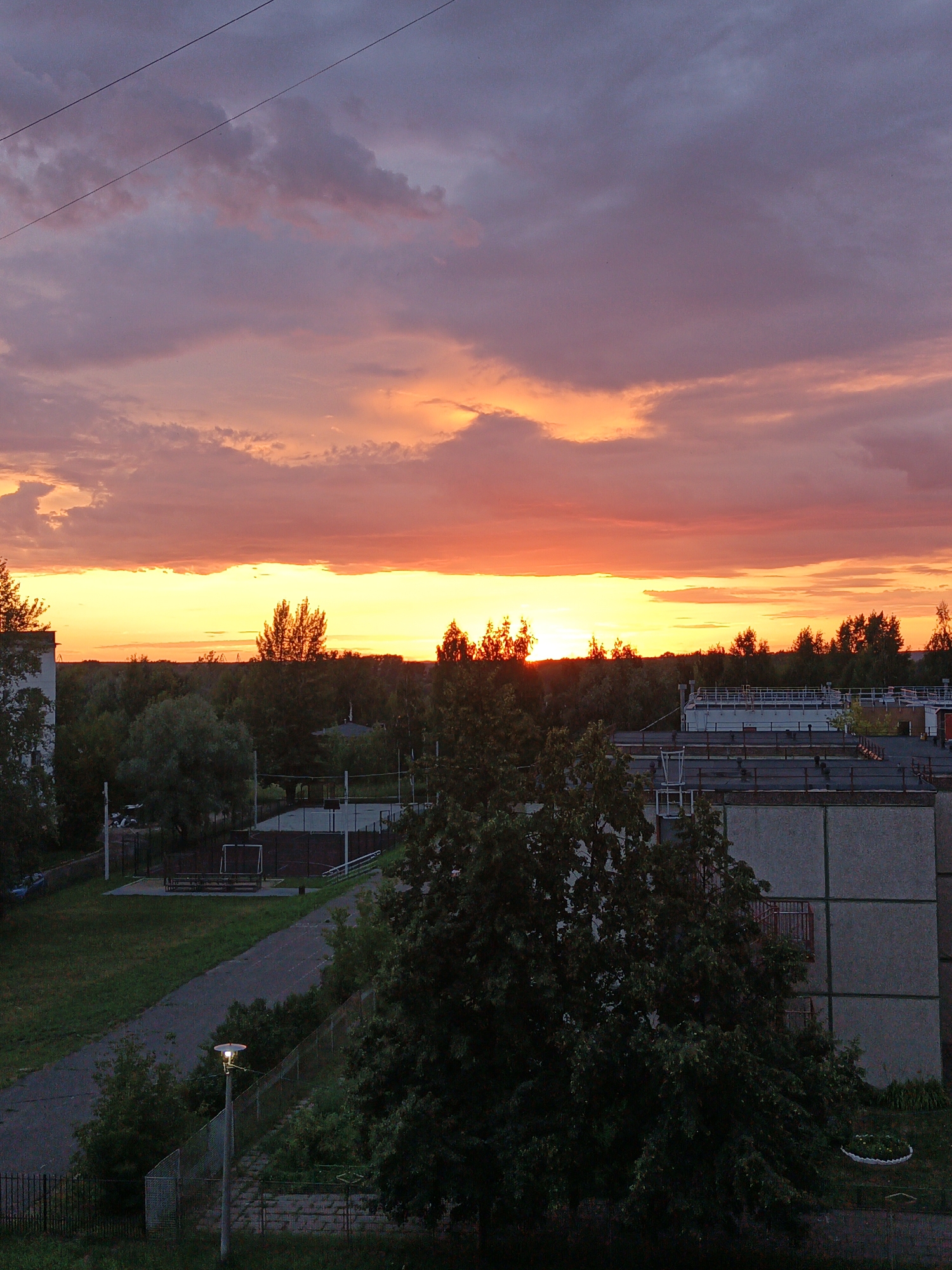 The image depicts a view of a residential area in Chaikovsky, Russia, during a sunset. The sky is a prominent feature, filled with vibrant hues of orange, yellow, and purple, indicating the setting sun. Clouds are scattered across the sky, some appearing wispy while others are more dense.
In the foreground and mid-ground, there are several elements of urban landscape. A multi-story building with concrete walls and visible windows is on the left side. In the center, a sports field with markings and a goalpost is visible, surrounded by a fence. Adjacent to the field is a paved path and a grassy area. A lone streetlight with a circular lamp is illuminating a section of the path. Trees, some appearing to be deciduous and others coniferous, form a dense line along the horizon, bordering the residential area and marking the edge of the settlement. Several other buildings, likely residential or industrial, are visible behind the trees. The overall scene is one of twilight in a suburban or semi-urban setting, characterized by the dramatic sky and the juxtaposition of natural elements with built structures. There are no people visible in the frame.