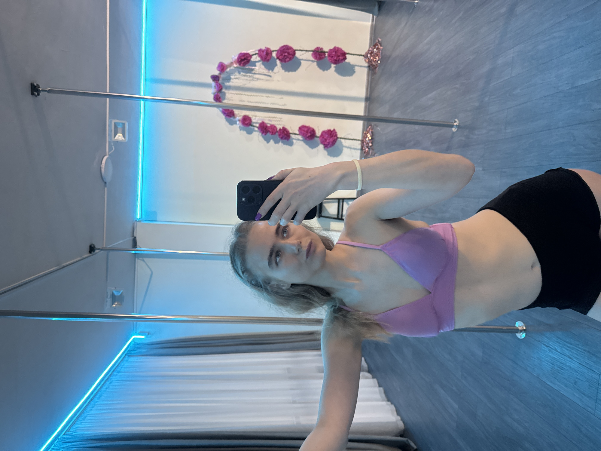A woman stands in front of a mirror, holding a smartphone to take a selfie in what appears to be a dance or fitness studio. She is wearing a purple sports bra and black shorts. The studio features a dance pole, blue neon lighting, and a wall decoration made of pink floral arrangements. The floor is bluish-grey.