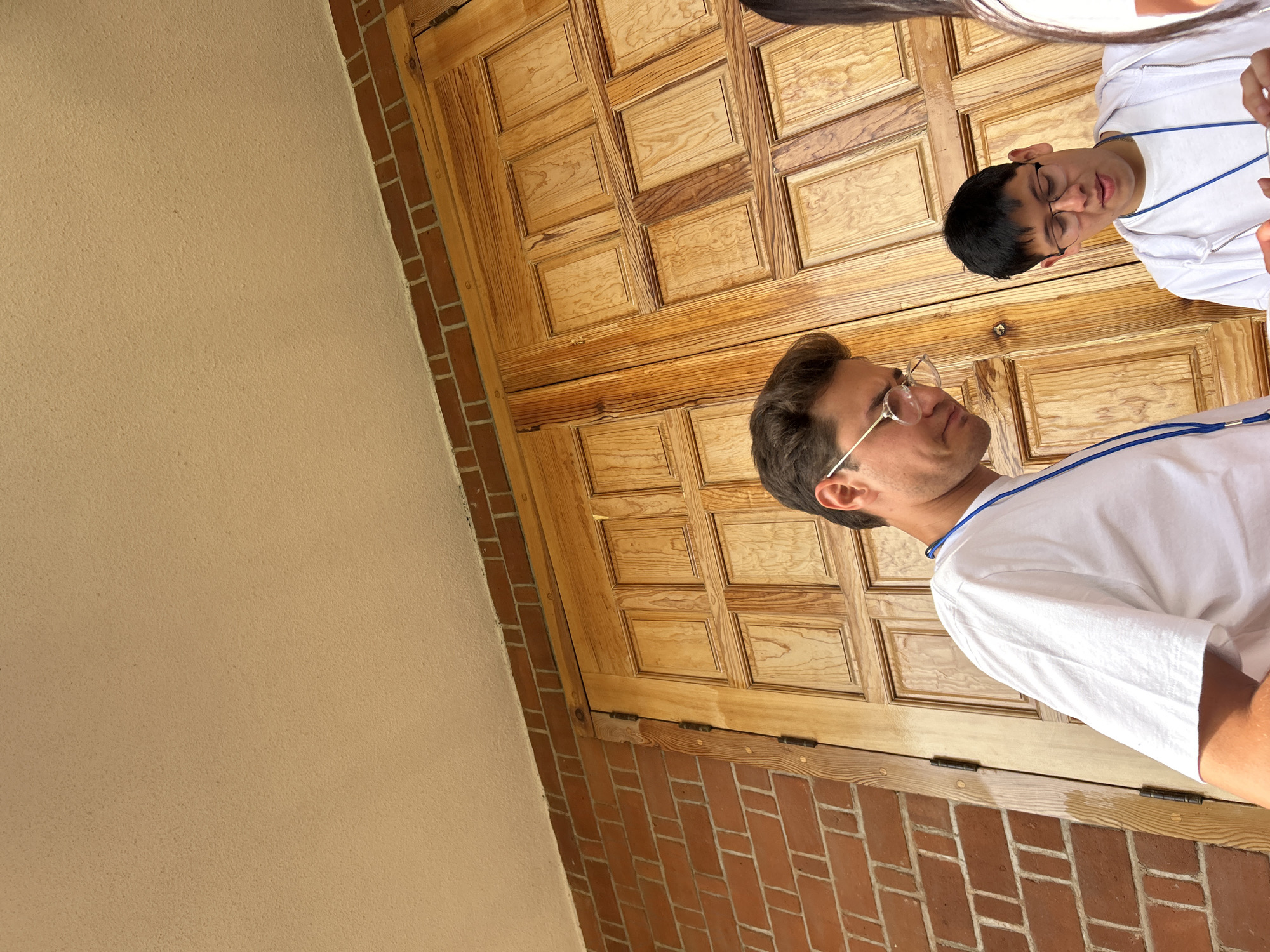 The image shows two individuals standing in front of a large, ornate wooden door set into a brick wall. The door is made up of multiple rectangular panels, and the brick wall appears to be part of a building's facade. To the left of the door is a light tan exterior wall. The two people are wearing white t-shirts with lanyards around their necks and appear to be engaged in a conversation or interaction. The man closest to the camera has glasses and a modern hairstyle, while the other person has shorter dark hair and also wears glasses. The location is Unknown, Unknown.