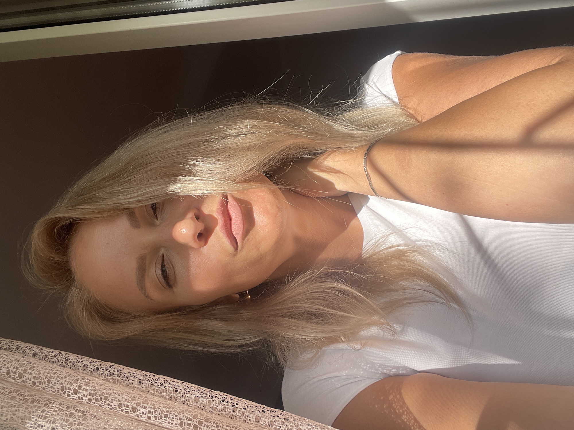 A woman with blonde hair wearing a white shirt is lying down with her head tilted back, her eyes closed, and a slight smile on her face. Her right hand is resting on her neck, and she is wearing a silver bracelet. The background consists of a dark wall or surface, a window frame, and a patterned fabric, possibly a curtain. Sunlight casts shadows across her face and clothing. There is no indication this is in Sochi, Russia.