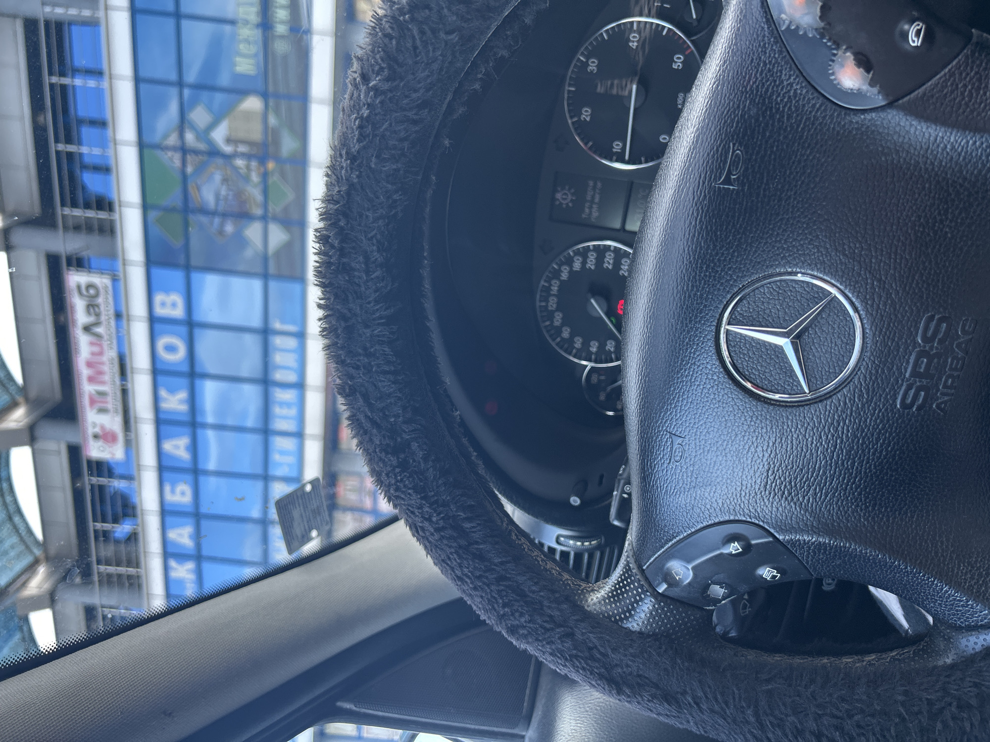 An image taken from inside a vehicle shows a fuzzy dark steering wheel with a Mercedes-Benz logo. Beyond the steering wheel, a car dashboard with a speedometer and tachometer is visible. Through the windshield, a building with blue windows and lettering that appears to be in Cyrillic script is seen. The context suggests the vehicle is in a city.