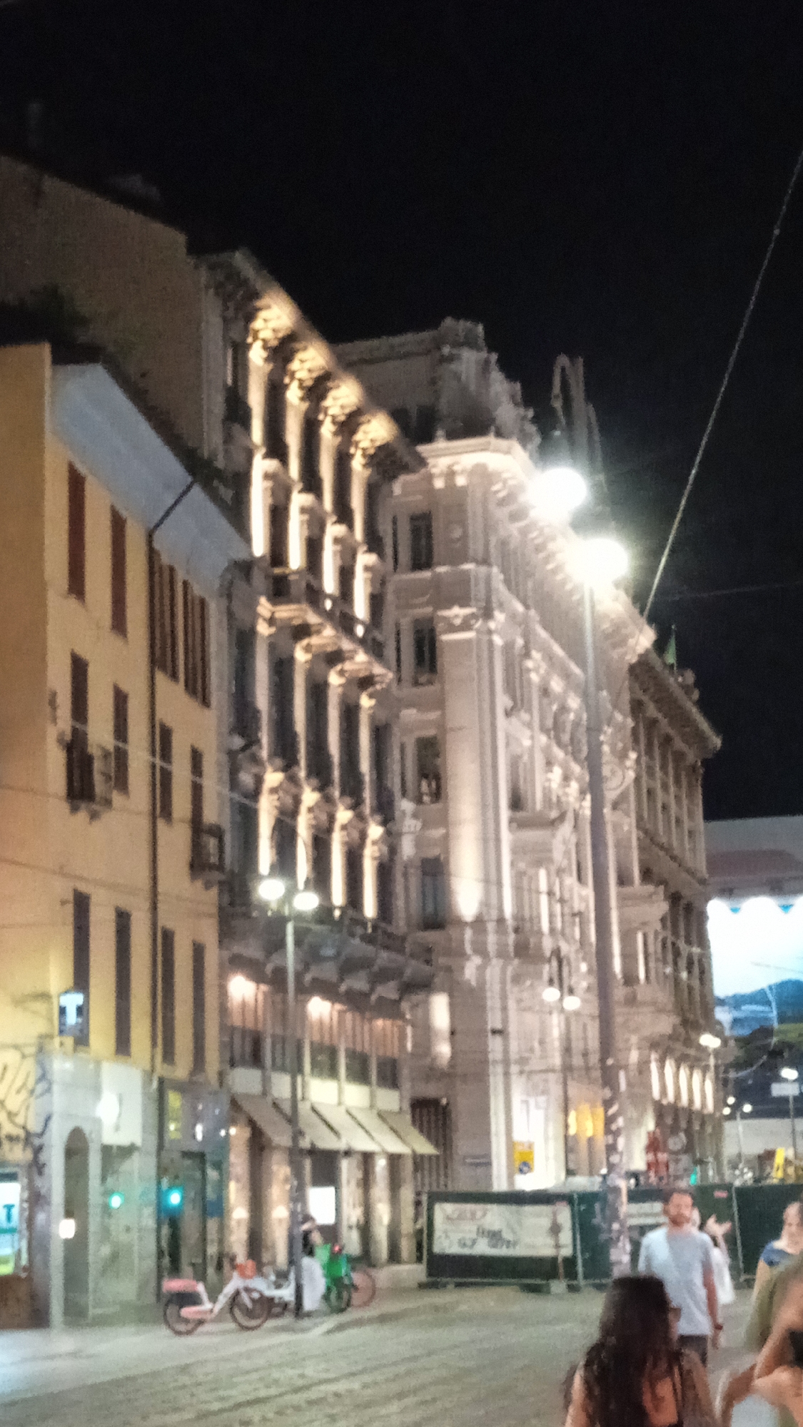 The image captures a street scene at night in Milan, Italy, likely in the city center. The architecture suggests an area with historical buildings, characterized by illuminated facades and decorative elements. The street appears to be paved and relatively wide, with some traffic and pedestrian areas.
In the foreground, a man in a light-colored t-shirt is walking towards the right. To his right, a woman with long dark hair is visible, also moving away from the camera. In the background, several buildings are illuminated by warm light, highlighting their architectural features like windows, balconies, and ornate detailing. Streetlights are also visible, contributing to the overall illumination of the scene.
On the left side of the image, there's a yellow building with dark-shuttered windows. Parked on the street are two bicycles, one with a pink seat and another green one. Signs and some construction barriers are also present, indicating ongoing urban activity. The sky is dark, typical of nighttime. The overall impression is a lively urban environment at night, with a mix of historical architecture and modern city life.