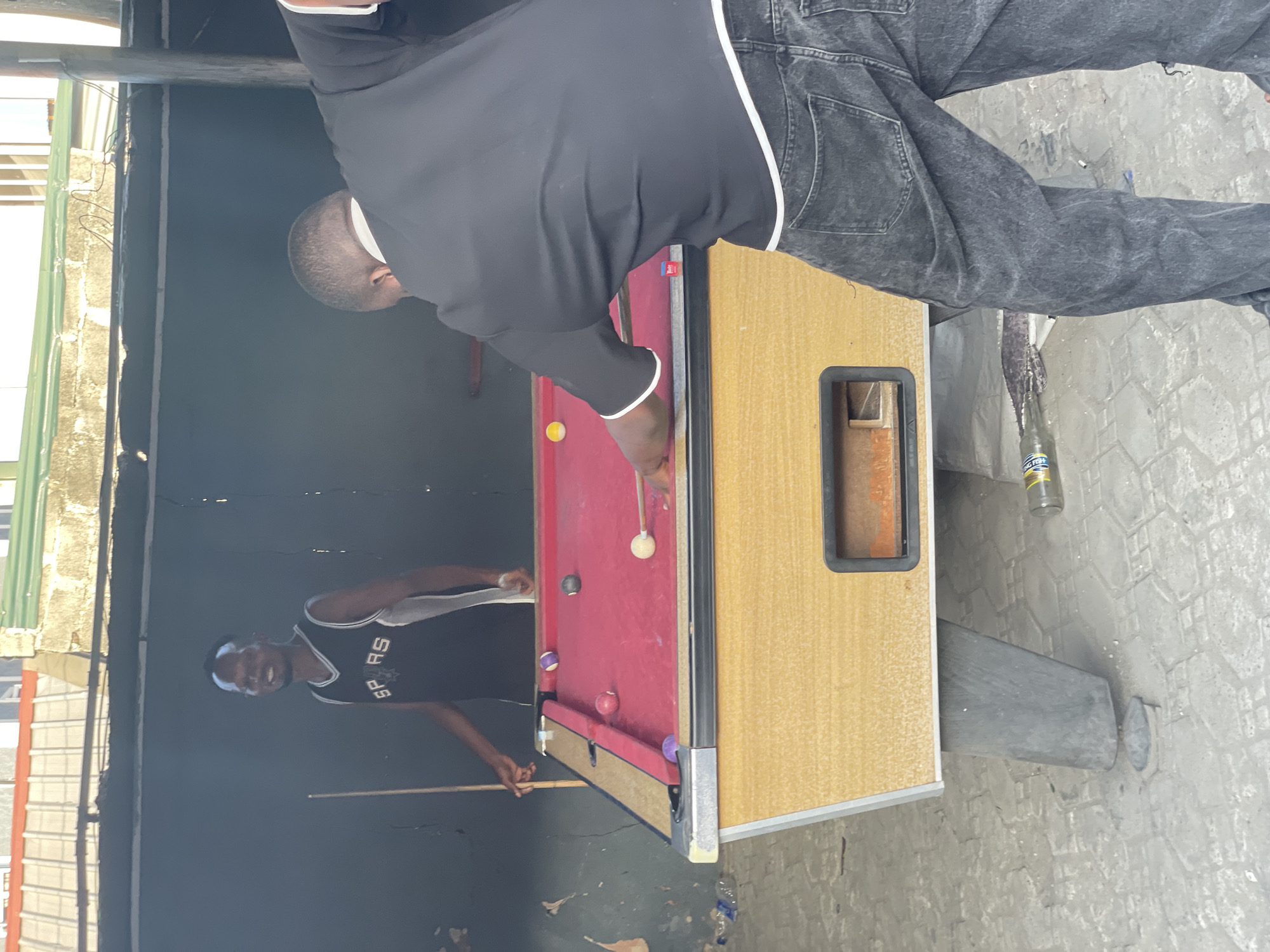 Two men play pool in an outdoor shed-like structure. One man, facing away, is bending over the pool table, cue in hand, dressed in a black top with a white stripe down the side and faded gray jeans. The pool table is red with tan sides. A second man, wearing a Spurs jersey, stands holding a cue stick. The structure has a tin roof, and the ground is paved. The shed is located in Ado, Nigeria.