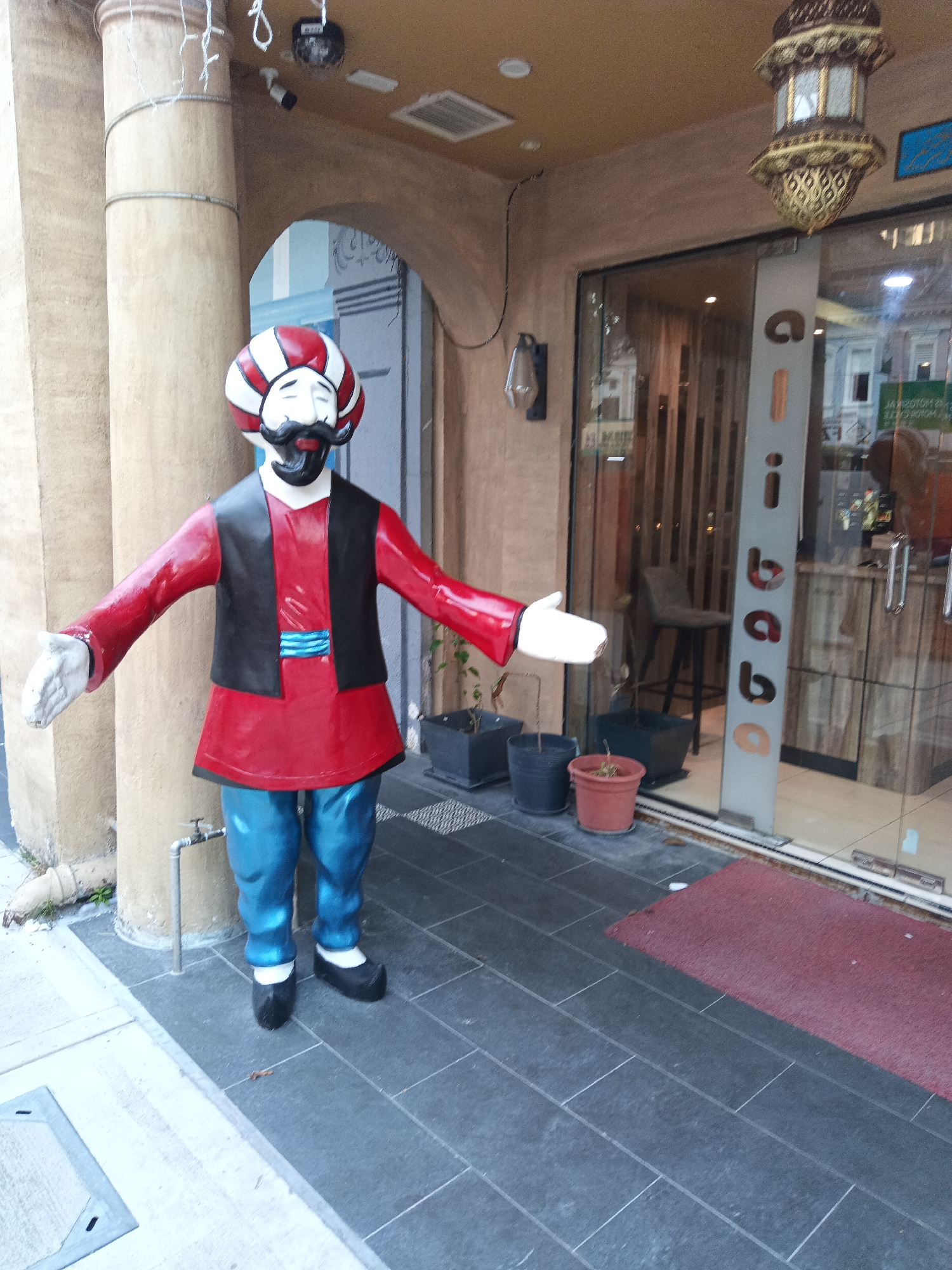 An animated statue of a man wearing a red tunic, blue pants, a black vest, and a red and white turban stands outside a shop in Kuala Lumpur, Malaysia. The statue's arms are outstretched. In the background, a glass-fronted shop displays items and interior seating. A decorative lamp hangs from the building's awning. The ground is paved with dark tiles, and a red mat is laid near the shop entrance.