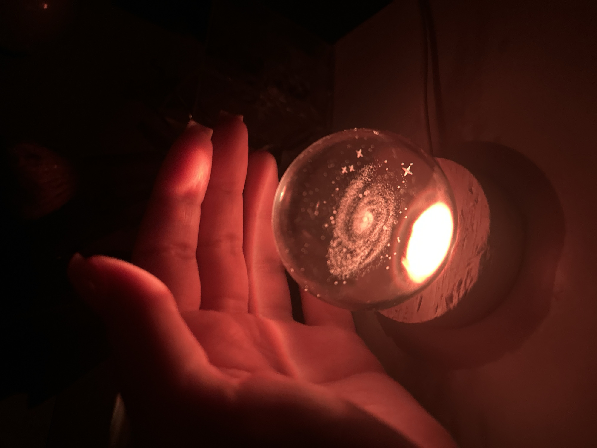The scene depicts a close-up, low-light view of a human hand interacting with a glowing spherical object in the city of Unknown, Unknown. A translucent glass sphere, approximately 4-6 inches in diameter, contains a three-dimensional representation of a spiral galaxy with numerous etched or suspended points of light, including several "X" shaped starbursts. The sphere rests on a textured, cylindrical base, from which a bright, warm-toned light source illuminates the sphere from below. A thin, light-colored cord extends horizontally from the right side of the base. A human hand, with skin appearing reddish due to the emitted light, is positioned on the left side of the frame, with fingers curved around or near the sphere. The immediate environment is dark, with the prominent red-orange glow from the sphere and its base casting reflections on the flat surface below.