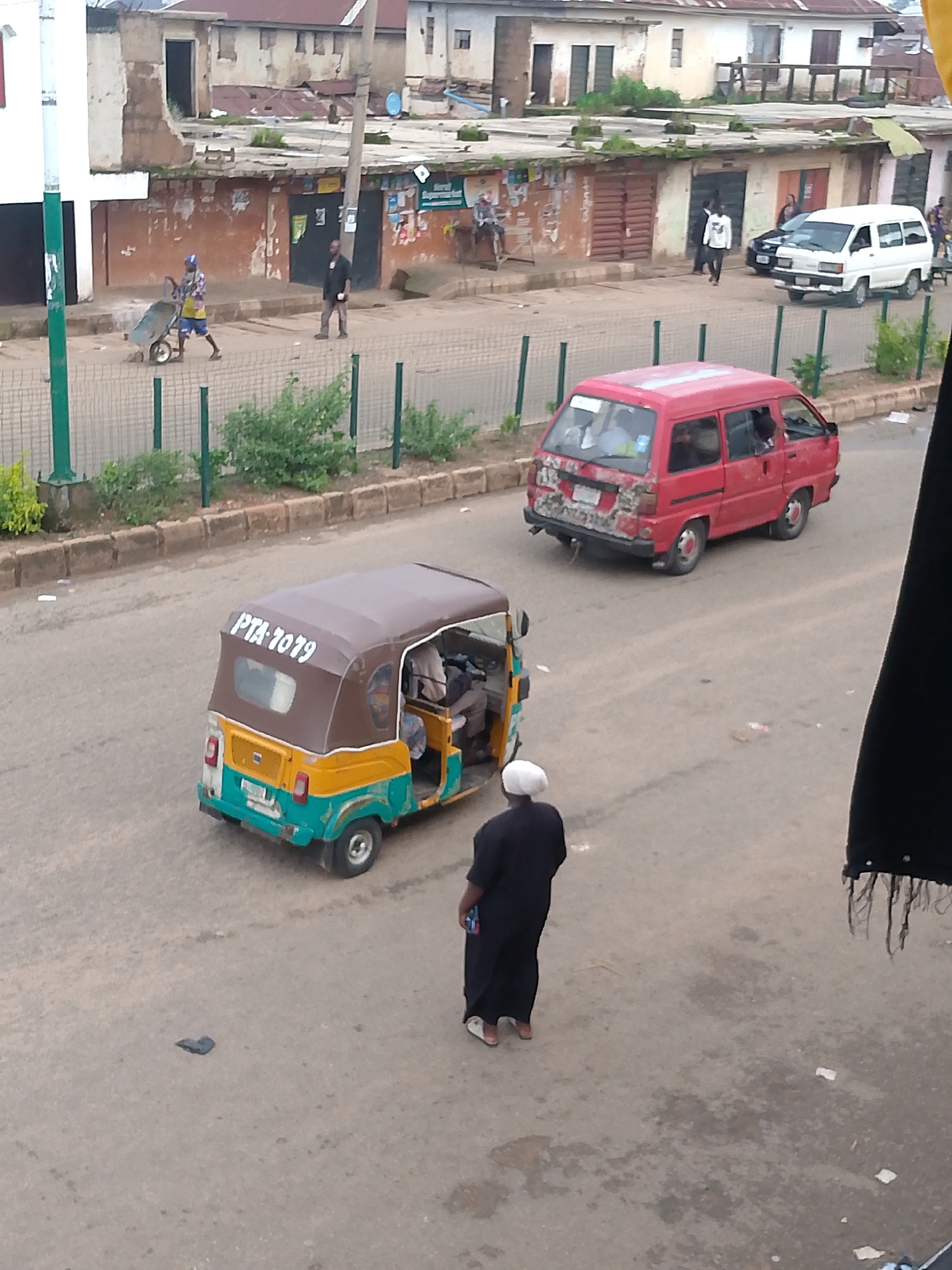 The image captures a street scene in Bukuru, Nigeria, with a focus on vehicular and pedestrian activity on a paved road. In the foreground, a woman dressed in a black abaya and white head covering stands facing away from the camera. To her left, a three-wheeled auto-rickshaw, predominantly yellow and teal with a brown canopy, is parked or moving slowly. The auto-rickshaw displays "PTA 7079" in white lettering on its canopy.
Further down the road, a red mini-van, appearing somewhat weathered with visible repairs on its rear, is in motion. The van has tinted windows, and the silhouettes of passengers can be discerned inside. Beyond the vehicles, a row of buildings forms the backdrop. These structures appear to be shops or residential dwellings, with some exhibiting signs of age and wear. A white mini-van is parked in front of one of these buildings.
In the upper left quadrant of the image, a person in a patterned shirt and blue shorts is pushing a wheelbarrow. This individual is on a sidewalk or pathway adjacent to the road, separated by a metal fence with green posts. Overgrown green vegetation is visible behind the fence. The overall environment suggests an active urban setting with a mix of old and new infrastructure. The lighting indicates it is daytime, likely overcast due to the soft shadows.