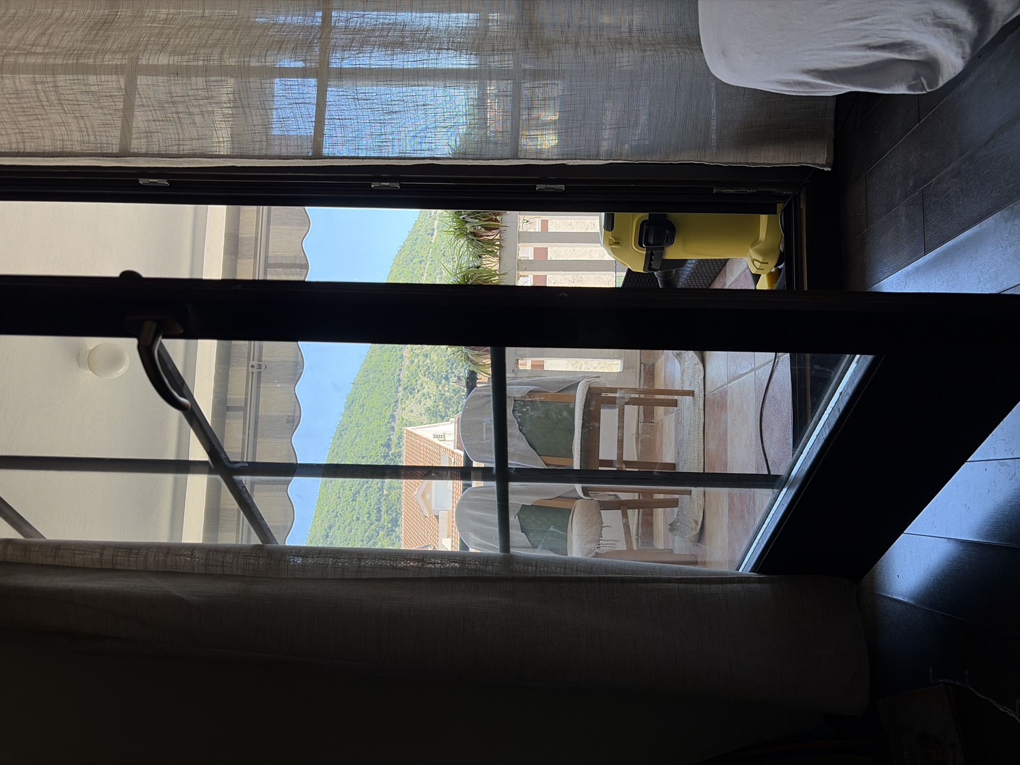 A view from inside a dimly lit room looking out through a window/door onto a balcony. The window/door frame is dark and partially obstructs the view. The balcony has chairs covered with cloths, and there is green vegetation visible. Beyond the balcony, a hill covered with green foliage is seen, along with rooftops of buildings. The sky is visible in patches. Inside the room, a curtain partially obscures the view. A vacuum cleaner can be seen on the balcony, and part of a bed is visible inside the room.
The location appears to be Budva, Montenegro, based on the architecture and the surrounding landscape.