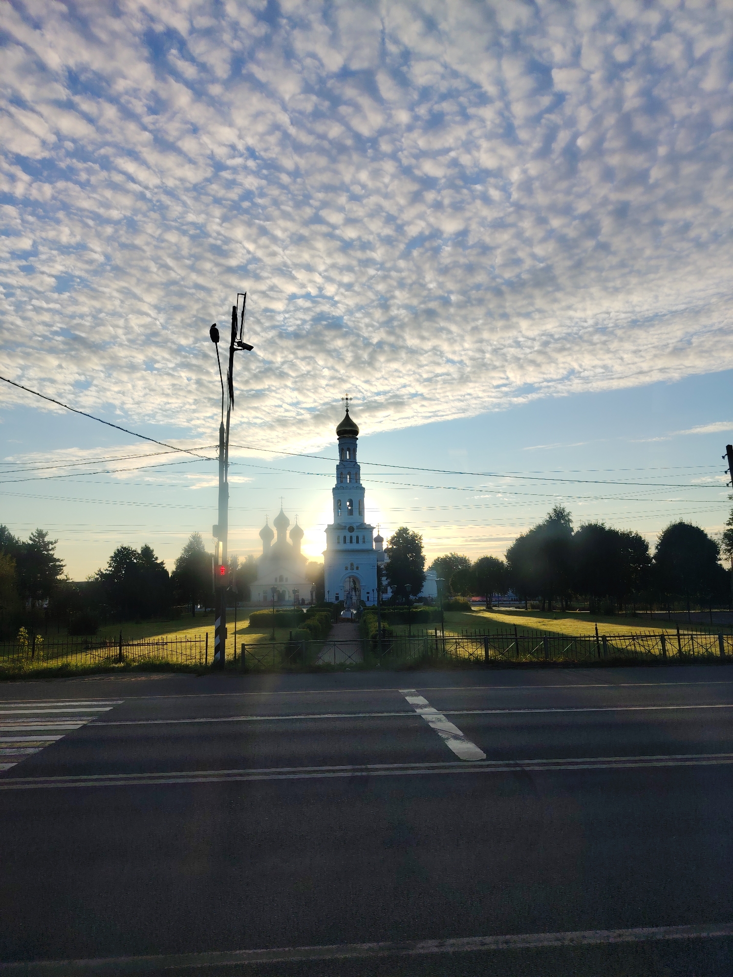 The image depicts the Ascension Cathedral in Zavidovo, Russia, at sunrise. The sky is filled with a dense pattern of stratocumulus clouds, illuminated by the rising sun which creates a bright glare behind the bell tower of the cathedral. The cathedral itself is white with a golden dome and a tall, white bell tower. In the foreground, a paved road with white lane markings and a pedestrian crossing is visible. A traffic light showing a red signal is mounted on a pole to the left of the road, with power lines strung across the frame. Beyond the road, a fenced-off grassy area leads to the cathedral, with trees lining the background. The overall scene is bathed in the warm, low light of early morning.