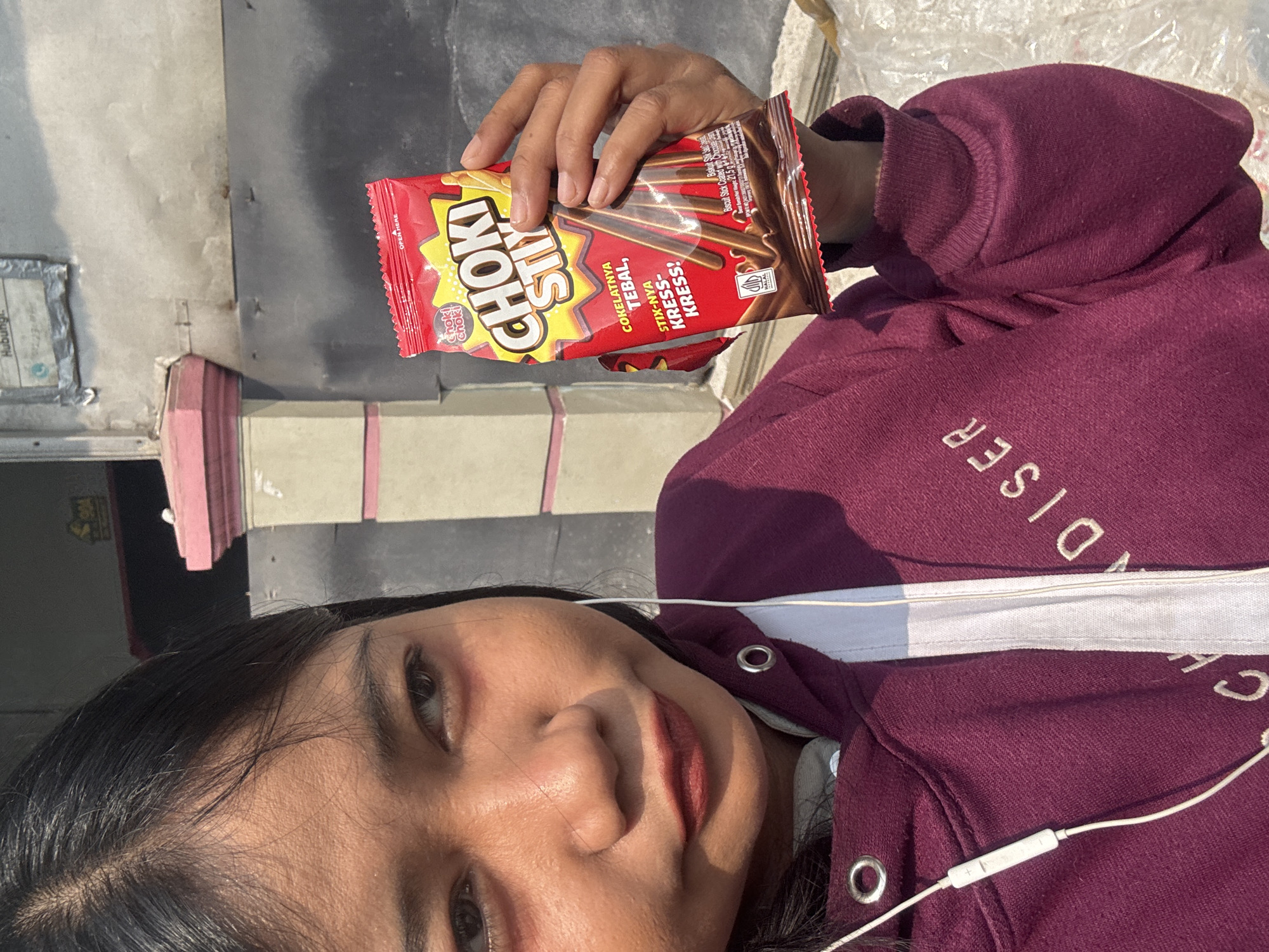 A young woman, appearing to be of Asian descent, is captured in a close-up, outdoor, selfie-style photograph. She has dark hair and is wearing a dark purple hooded sweatshirt. She is holding a red package of "Choki Stix" chocolate biscuits in her right hand. The packaging features a yellow starburst design and images of chocolate-covered biscuit sticks. The woman is looking off to the left of the frame. Her left ear is adorned with a white earbud connected to a white cord that trails down her sweatshirt.
The background suggests an urban setting in North Jakarta, Indonesia. A weathered, grey metallic wall is visible behind the woman, with some visible rust and a sign attached to it. To the left of the wall, a portion of a pink and cream-colored architectural structure, possibly a pillar or decorative element, is present. The lighting indicates it is daytime, with sunlight casting shadows and highlighting the texture of the woman's skin and the objects in the frame. A plastic-wrapped object, possibly merchandise or packaging, is partially visible on the right side of the image. The overall scene suggests a casual moment captured outdoors, likely on a street or in a commercial area within North Jakarta.