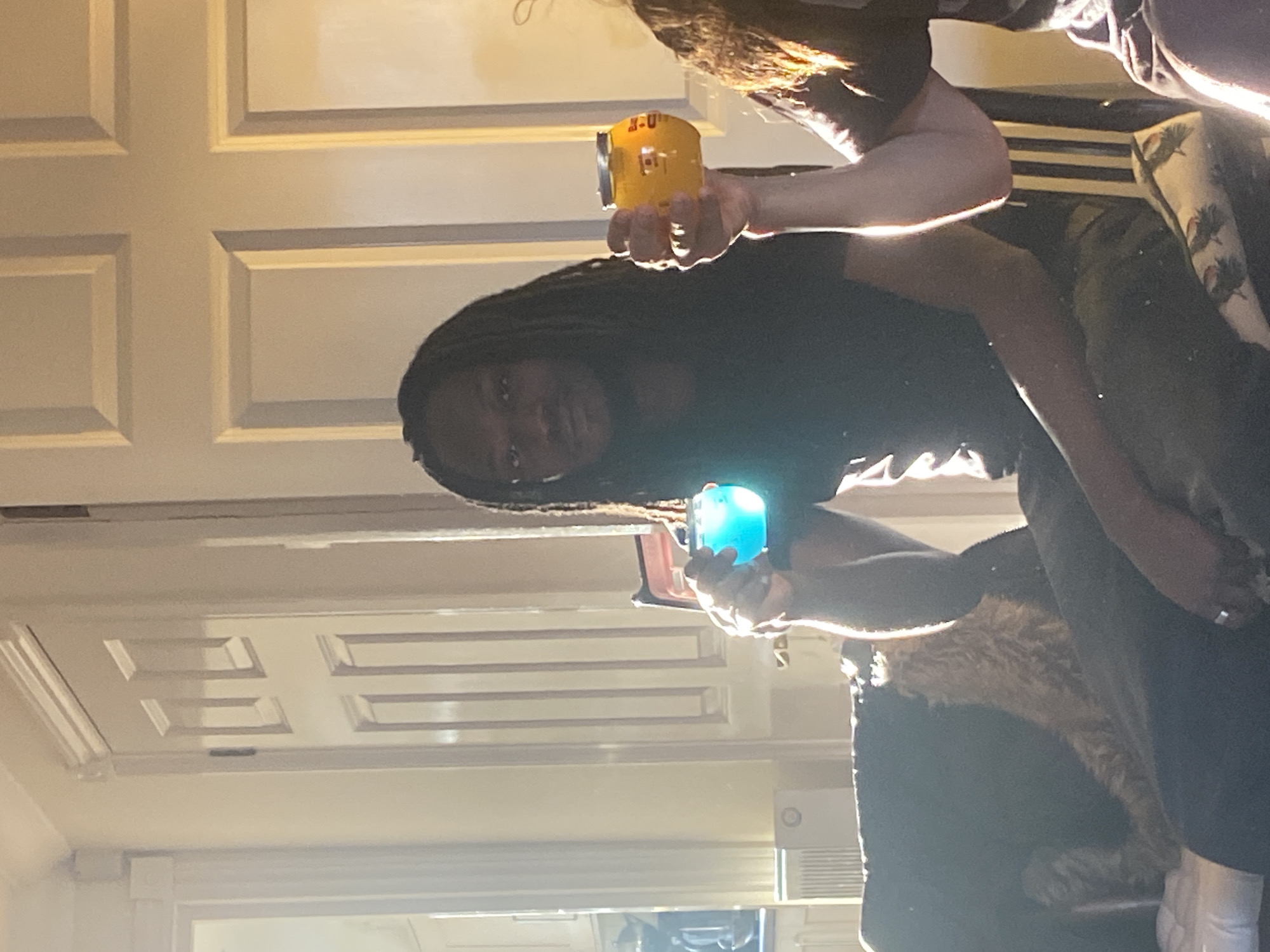 A person with dreadlocks is holding a yellow can in their right hand and a blue can in their left hand. The person is wearing a black shirt. Another person is partially visible in the top right of the image, wearing a striped shirt. The setting appears to be indoors, with a door visible in the background. I have no information from the image to determine the location in Reigate and Banstead, United Kingdom.