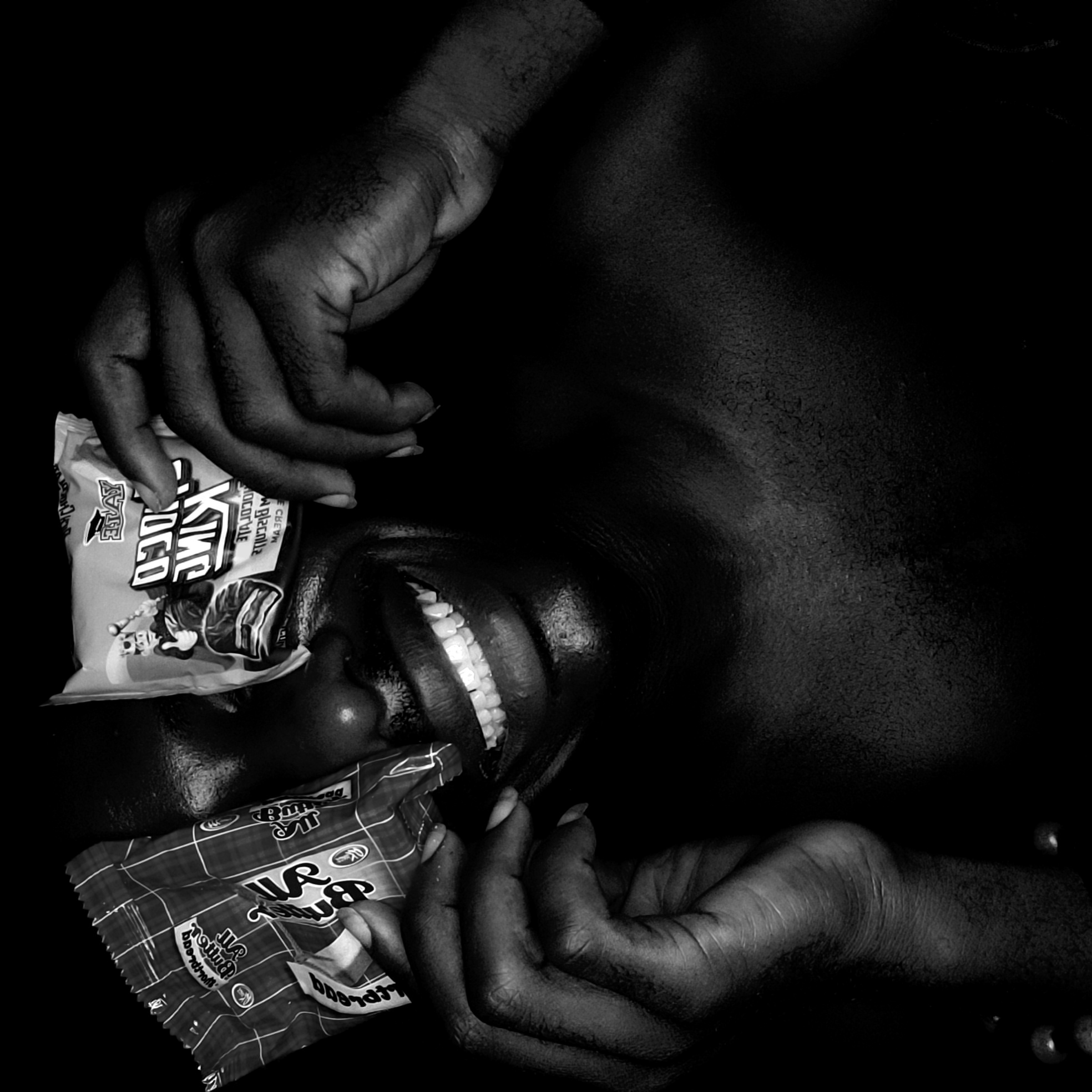 A monochrome photograph depicts a person lying with their head tilted back. A snack bag, "King Choco" rests on their forehead. Another snack bag is held near their mouth with one hand. The person's mouth is open in a wide smile, revealing their teeth. The person's body is partially visible, set against a dark background. The location of Narayi, Nigeria is not discernible from the image.