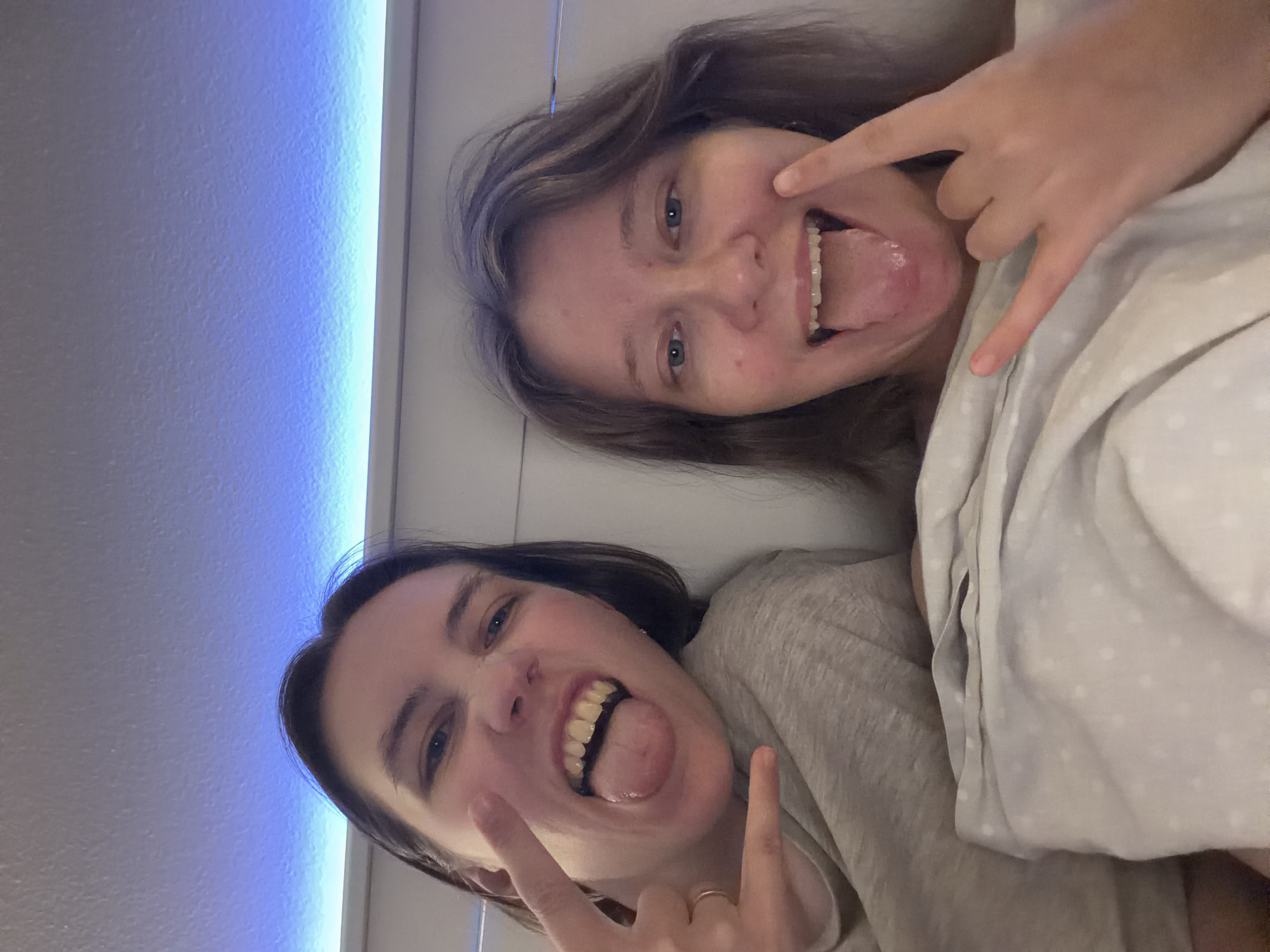Two young women are lying in bed, taking a selfie. They are both sticking out their tongues and making "rock on" hand gestures. The woman on the left has dark hair and is wearing a gray shirt. The woman on the right has lighter brown hair and is wearing a white shirt. They are covered by a light-colored blanket with a white polka dot pattern. The background shows a wall with blue LED lighting. The setting appears to be a bedroom.