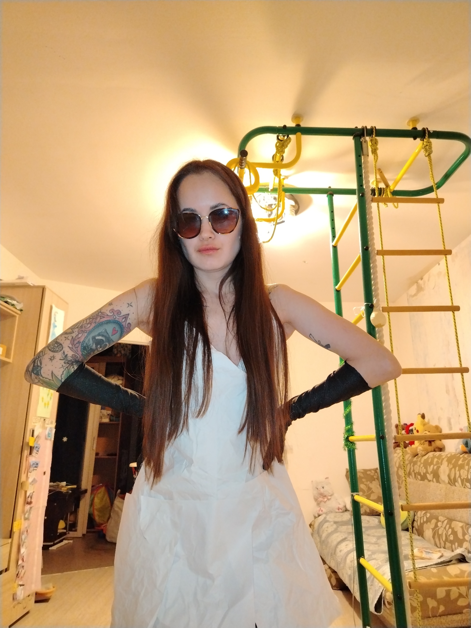 A young woman with long, straight, dark brown hair is posing indoors. She is wearing sunglasses, a white crinkled-fabric dress, and long black arm coverings or fingerless gloves. Tattoos are visible on her forearms; her left arm features a colorful tattoo with a raccoon in a heart. She stands with her hands on her hips, looking at the camera with a neutral expression.
The setting appears to be a residential room, possibly in Noviy, Russia, characterized by an indoor children's play structure with green metal bars, yellow rungs, ropes, and rings, which dominates the right side of the frame. A sofa with a floral or leaf pattern is situated behind the play structure, decorated with several stuffed animals, including teddy bears and a white cat plush. On the left, part of a light-colored wooden cabinet is visible, adorned with various papers or photos held by magnets. The room has light-colored walls, and ceiling lights are on, suggesting it is either daytime indoors or evening. No visible text is present in the image.