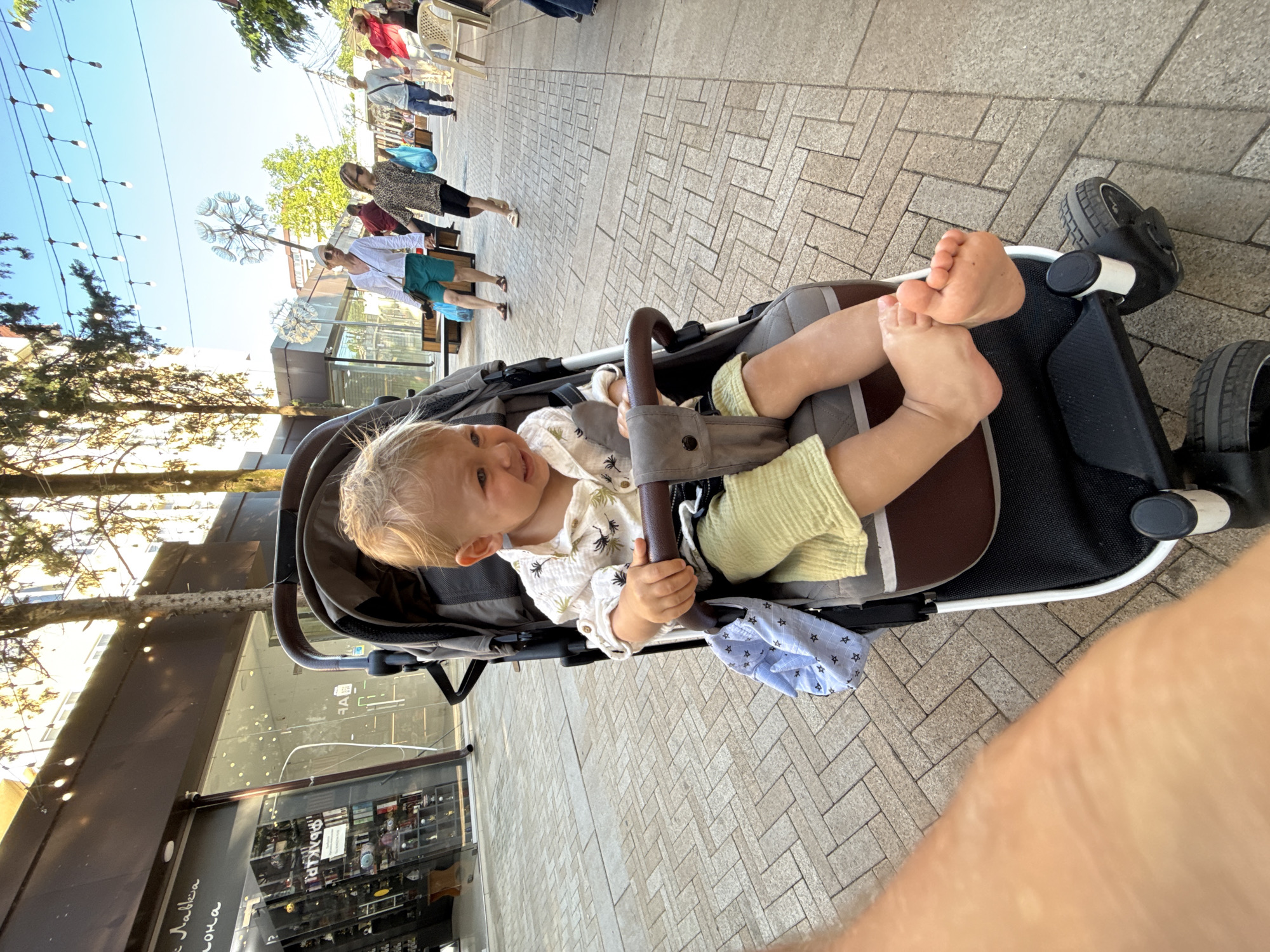 A young child with blonde hair sits in a stroller on a paved walkway lined with shops. The child wears a light-colored shirt with a palm leaf pattern and light green shorts, and is barefoot. The stroller is dark gray with a brown handle and has a blue cloth bag hanging from it. In the background, people walk along the pathway under strings of lights. Trees are visible, and the sky is clear and blue. The location is identified as Sirius, Russia.