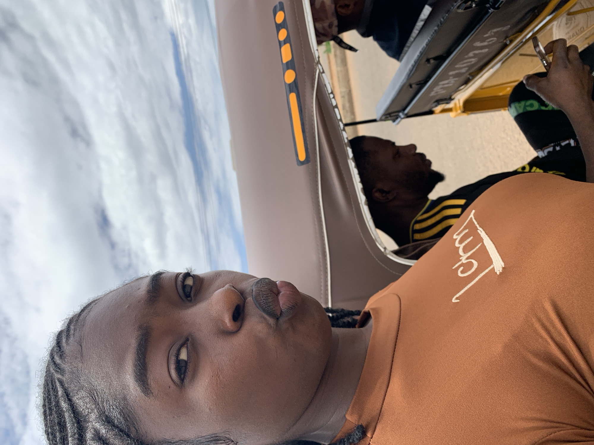 The image captures a selfie-style perspective from inside a motor tricycle (Keke Napep) in Rafin Par, Nigeria, under an overcast sky.
In the foreground, a dark-skinned woman with braided hair looks directly at the camera, her lips pursed in a pout. She wears a rust-colored short-sleeved top with white stylized embroidery on the left chest, appearing to read "Lctm". Her right arm is visible, extended towards the camera.
Behind her, to the right from the viewer's perspective, a dark-skinned man with a beard is visible in profile, looking down at a silver-colored smartphone held in his right hand. He wears a black shirt with yellow horizontal stripes, possibly a sports jersey, and dark pants or shorts with green lettering visible on his left leg.
Further back and to the right, only the top of another individual's head, wearing a dark cap, is partially visible.
The vehicle's interior features a brown canopy roof with white trim, and a black rectangular reflector strip with orange circular and rectangular elements affixed to its underside. A black backrest or storage area, bearing partial white numbering "PPA 1076", is visible behind the man holding the phone. The vehicle's frame appears to be yellow. The background shows a blurry road or ground with a reddish-brown hue and an expansive cloudy sky.