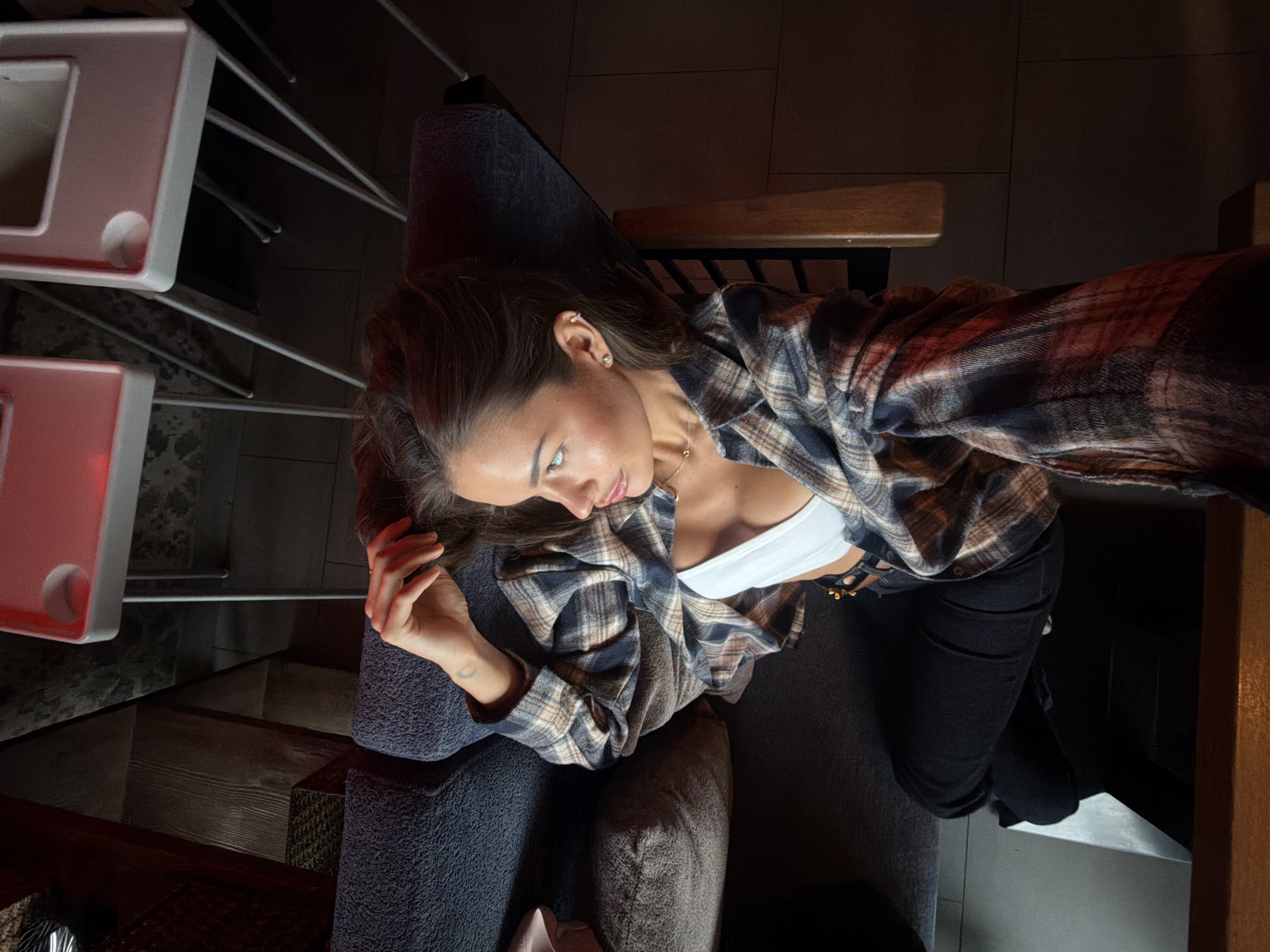 A young woman with long, dark brown hair is lying on a dark blue sofa, viewed from above. She is wearing a white bralette, black pants, and an open, plaid shirt. Her right arm is raised, with her hand resting on her head, and her left hand is down by her side. She is looking to her right, with her profile visible. She has a small necklace around her neck and a visible ear piercing. Behind her, two white tables with pink accents are visible. The overall lighting suggests an indoor setting, possibly a lounge or cafe, though no specific location in Krasnoyarsk, Russia can be determined from the image.