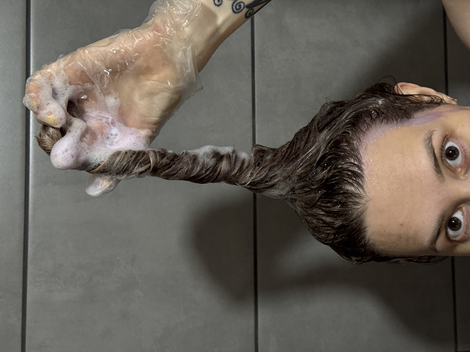 A person with light skin and brown hair, possibly female, is having their hair washed. The hair is being held by a gloved hand covered in soapy lather. The person has visible brown eyes and is looking upwards. The background is a gray tiled wall, typical of a bathroom. There is no information to indicate the location is Chaikovsky, Russia.
