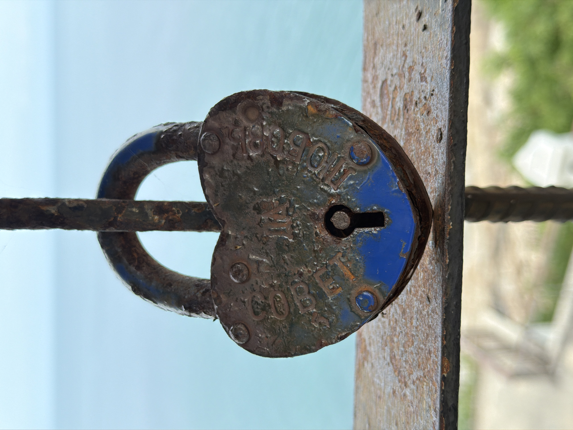An old, heart-shaped, rusted padlock hangs on a metal railing, obscuring the view of the ocean in the background. The padlock is weathered and shows signs of blue paint chipping away. Raised lettering on the padlock is partially visible, with the words "COBET" and "LONDON" discernible. The railing it hangs on is also rusty and appears to be part of a fence or balcony. In the blurred background, a body of water, likely the sea, is visible under a light sky. Below the railing, the scene is out of focus, showing hints of greenery and structures, suggesting an outdoor setting, possibly in a coastal area. There are no people or animals present in the image. The lighting suggests it is daytime.