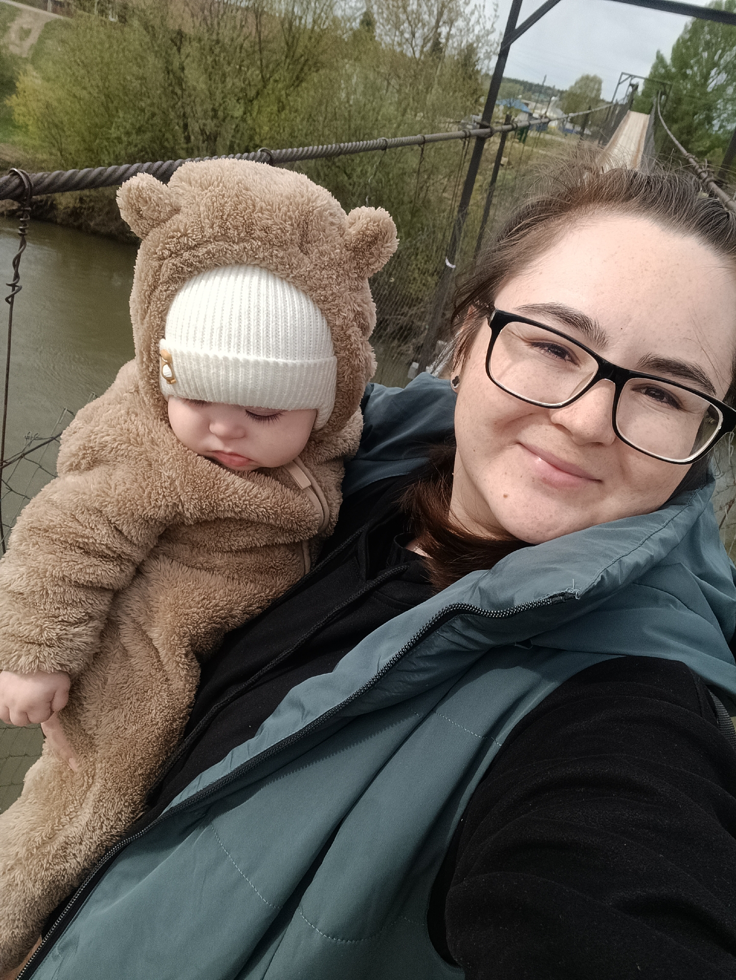 A woman holds a baby on a suspension bridge in the city of Посад, Russia. The woman, wearing glasses and a blue vest, smiles at the camera. The baby, clad in a brown bear-like snowsuit and a white beanie, has their eyes closed and appears to be sleeping. The suspension bridge, with its cables and wooden planks, spans over a river. Trees and buildings are visible in the background.