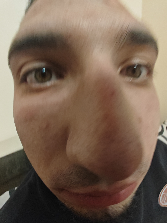 A close-up, wide-angle photograph depicts an adult male's face, significantly distorted by lens effects, with the nose centered and disproportionately enlarged. The subject exhibits light skin, light brown eyes with prominent vertical reflections, and dark eyebrows. Fine stubble is visible on the upper lip and chin. The individual is wearing a dark-colored shirt, with a light-colored, circular emblem featuring a reddish-orange design visible on the right side. The background consists of an out-of-focus indoor setting, characterized by light-toned walls and an indistinct dark object to the left. No identifiable landmarks or specific geographical indicators are present to determine a location within Varna, Bulgaria, or any other city.