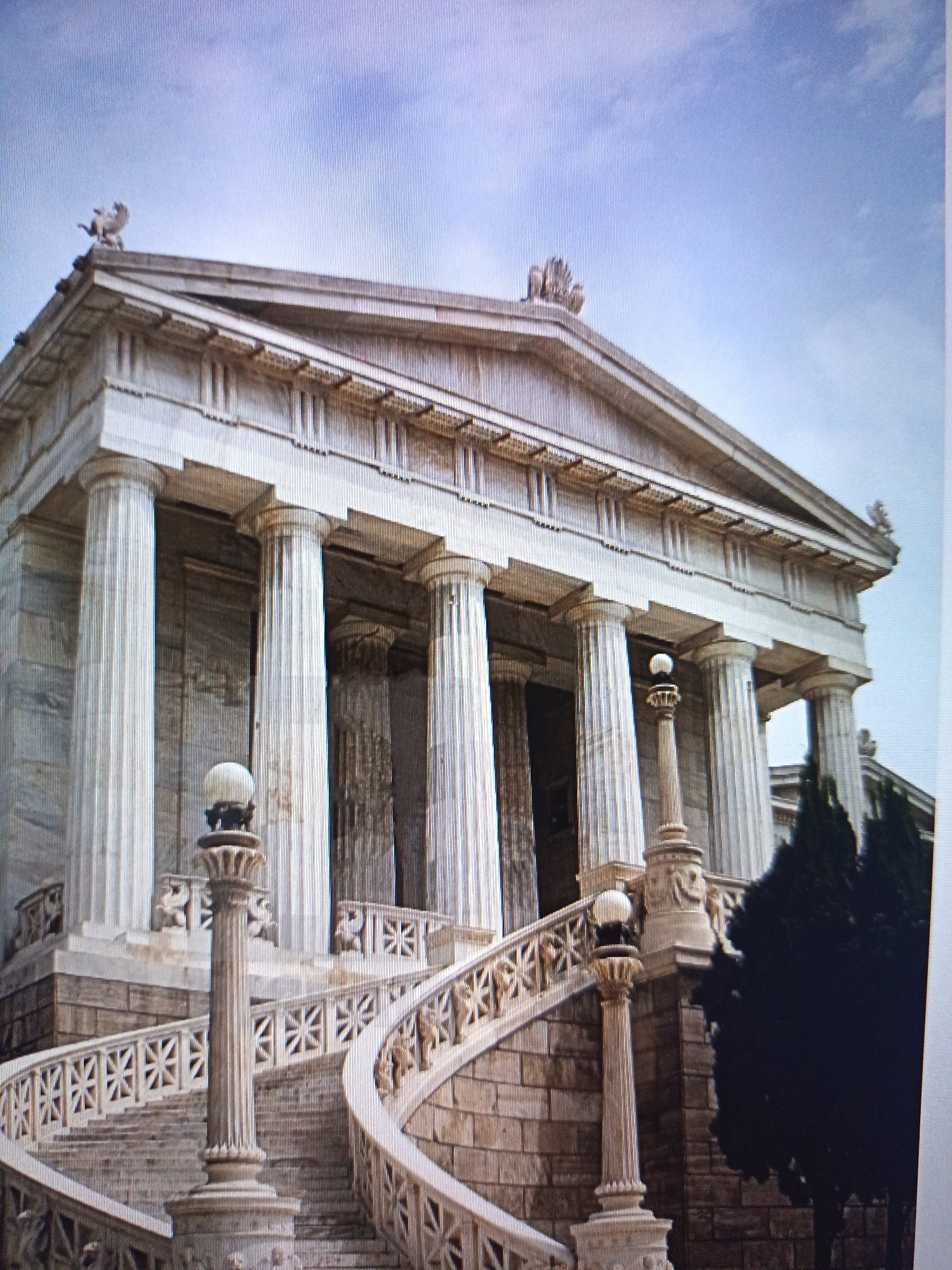 The image shows a grand neoclassical building with a pedimented facade, supported by multiple fluted columns. A curved staircase with ornate railings leads up to the building's entrance. Sculptural elements, possibly lions, are visible on the roofline. A tall column with a globe-like fixture stands beside the staircase. A dark evergreen tree is situated to the right of the building. The sky above is a pale blue with some scattered clouds. This building is the National Library of Uzbekistan named after Alisher Navoi, located in Tashkent, Uzbekistan.
