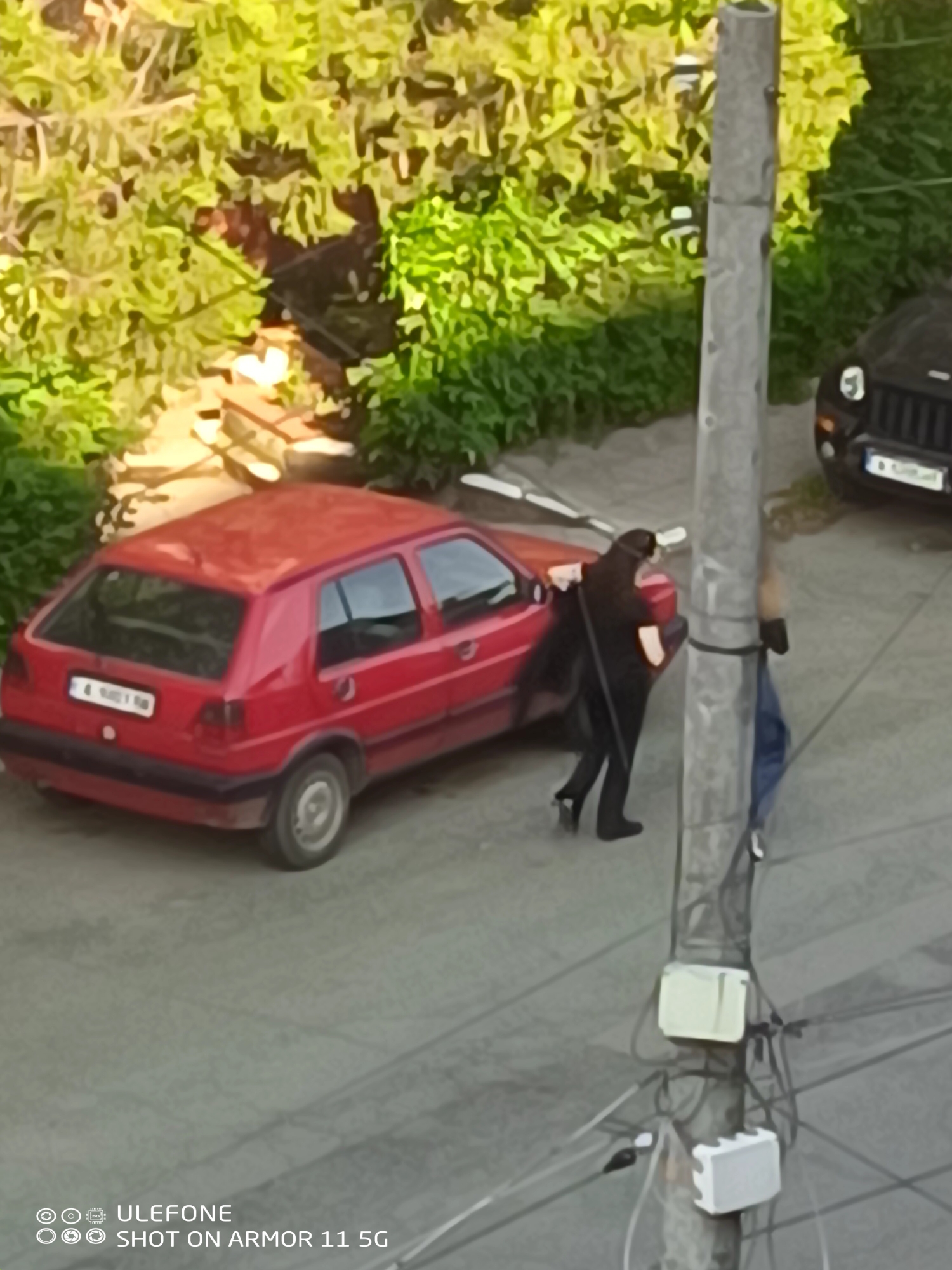 The image captures an elevated view of an outdoor scene during daytime in Unknown, Unknown, featuring two individuals, two vehicles, and surrounding greenery.
**People:**
1. One individual is observed on the driver's side of the red hatchback car. This person is dressed in dark attire, including what appears to be a long coat or dress and dark shoes with discernible heels. Their posture suggests they are either entering or exiting the vehicle. A dark head covering or hair is visible.
2. A second individual is partially visible on the right side of the frame, largely obscured by a utility pole. This person is wearing blue pants, consistent with denim, and a light-colored upper garment. They appear to be standing beside the pole.
**Vehicles:**
1. A red two-door hatchback car, consistent with a Volkswagen Golf Mk2 or Mk3 model, is centrally parked with its rear facing the viewer. The rear license plate is partially legible, displaying "4 W98116". The vehicle exhibits signs of external wear and dirt.
2. The front section of a black SUV or Jeep-type vehicle is partially visible on the far right, positioned perpendicular to the red car. Its front grille and a portion of the hood are discernible, along with an unreadable front license plate.
**Environment and Objects:**
The ground is a paved surface, likely asphalt or concrete. Dense green foliage, comprising trees and bushes, occupies the background and the left side of the frame, bordering the paved area. A prominent grey utility pole stands in the mid-right foreground, featuring multiple attached dark cables and two rectangular electrical boxes, one cream-colored and one white, mounted vertically. Behind the red car, amidst the greenery, low structures made of light-colored stone or brick are visible, possibly forming steps or a wall.