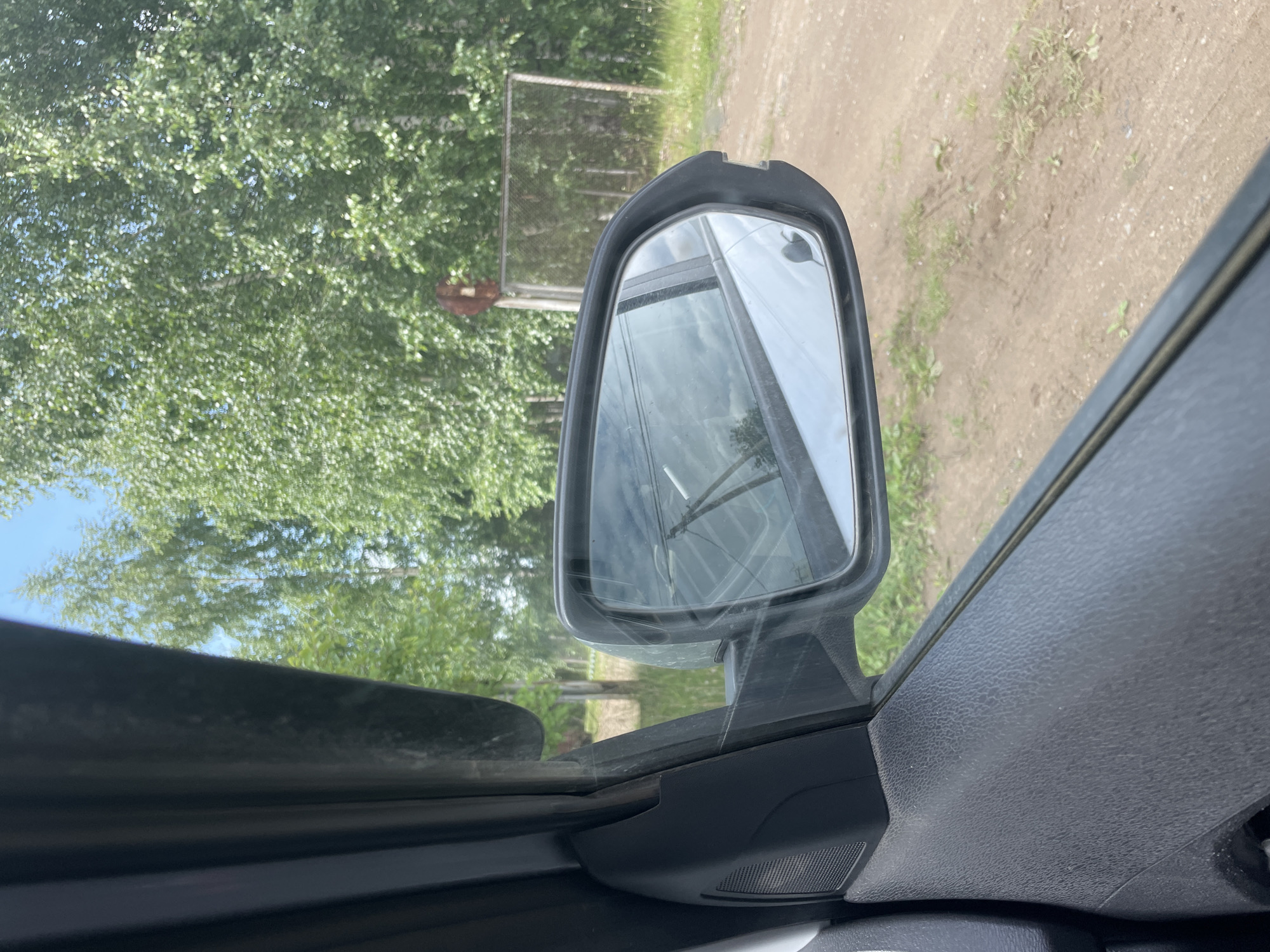 The media file is a first-person perspective photograph taken from inside a vehicle, looking out through the side window and mirror. The scene is outdoors in a rural or semi-rural setting.
The primary subjects are the car's interior elements, specifically the side mirror housing and a portion of the car door panel, which are made of black textured plastic. The side mirror itself is visible, reflecting the surroundings. Outside the car, the view is dominated by lush green trees and foliage, suggesting a time of year with abundant plant growth, likely spring or summer. A bright blue sky is visible through the leaves, indicating fair weather.
In the reflection of the side mirror, a metal fence or gate is partially visible, along with a dusty dirt road and more trees. There is also a hint of a cloudy sky in the reflection. The overall impression is of a sunny day, possibly late morning or early afternoon, given the lighting. The car appears to be parked or slowly moving near a treeline next to a dirt road. There are no people or animals clearly visible in the frame.
Notable details include the texture of the car's interior plastic and the clarity of the reflection in the side mirror, which allows for a clear view of the exterior scene. The image provides a sense of being on a journey or a drive through a natural landscape. No visible text is present in the image. The location context is specified as Nozhovka, Russia.