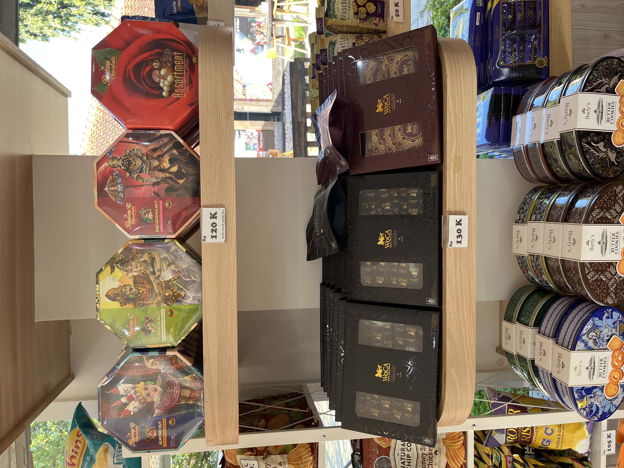 The image depicts a retail shelf in a store selling various food items, likely in Unknown, Indonesia, given the presence of Indonesian Rupiah (Rp) price tags and the brand "Baline Chocolate." The shelf is divided into sections, each displaying different products.
On the left side are stacks of hexagonal boxes of Baline Chocolate in different flavors. From top to bottom, there's "Assortment," "Macadamia Nut," "Cashew Nut," and "Almond Nut." A price tag of Rp 120K is visible next to these items.
In the middle section, there are stacks of "WOCA" brand chocolate boxes. The boxes are mostly black with gold lettering, although there are also some brown and gold colored boxes. A price tag of Rp 130K is placed next to these.
On the right side are stacks of "Barly's Butter Cookies" in round tins, various designs. Below these cookie tins are bags of "The Natural Chip Co," "Maretti Bruschetta Chips," and "Rp. Hand Cooked Chips".
The environment appears to be a well-lit store, with glimpses of the outdoors visible in the background. The overall impression is a well-stocked shelf with a variety of sweets and snacks for sale.