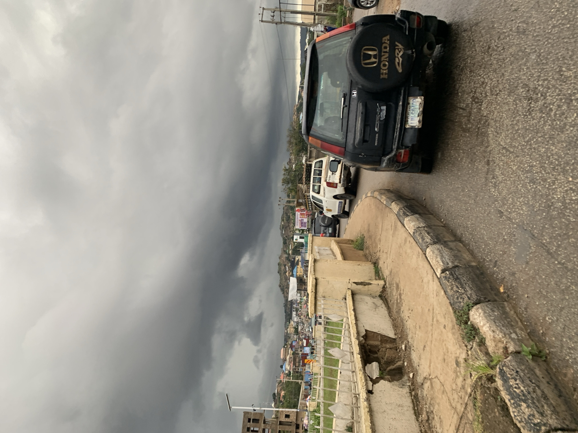 The image captures a street scene in Jos, Nigeria, under a heavily overcast sky. The foreground features a road with a curb on the left, which appears worn and cracked, with some vegetation growing out of it. A black Honda CRV is parked on the right side of the road, facing away from the viewer. Its spare tire, mounted on the back, has a Honda logo and "CRV" text visible. A license plate with the text "NLM 594DD" is affixed to the rear of the vehicle. To the left of the Honda CRV, a white pickup truck is visible in traffic.
Further in the background, there is a cityscape with buildings of varying sizes and colors spread across rolling hills. Some buildings appear to be under construction or in a state of disrepair. A white metal fence with diamond-shaped openings runs along the side of the road in the middle ground, adjacent to a beige concrete structure that is also showing signs of damage and wear. Traffic is visible on the road, suggesting a bustling urban environment. The overall atmosphere is somber due to the dark, cloudy sky, which hints at impending rain. The lighting is diffused, creating a muted color palette across the scene.