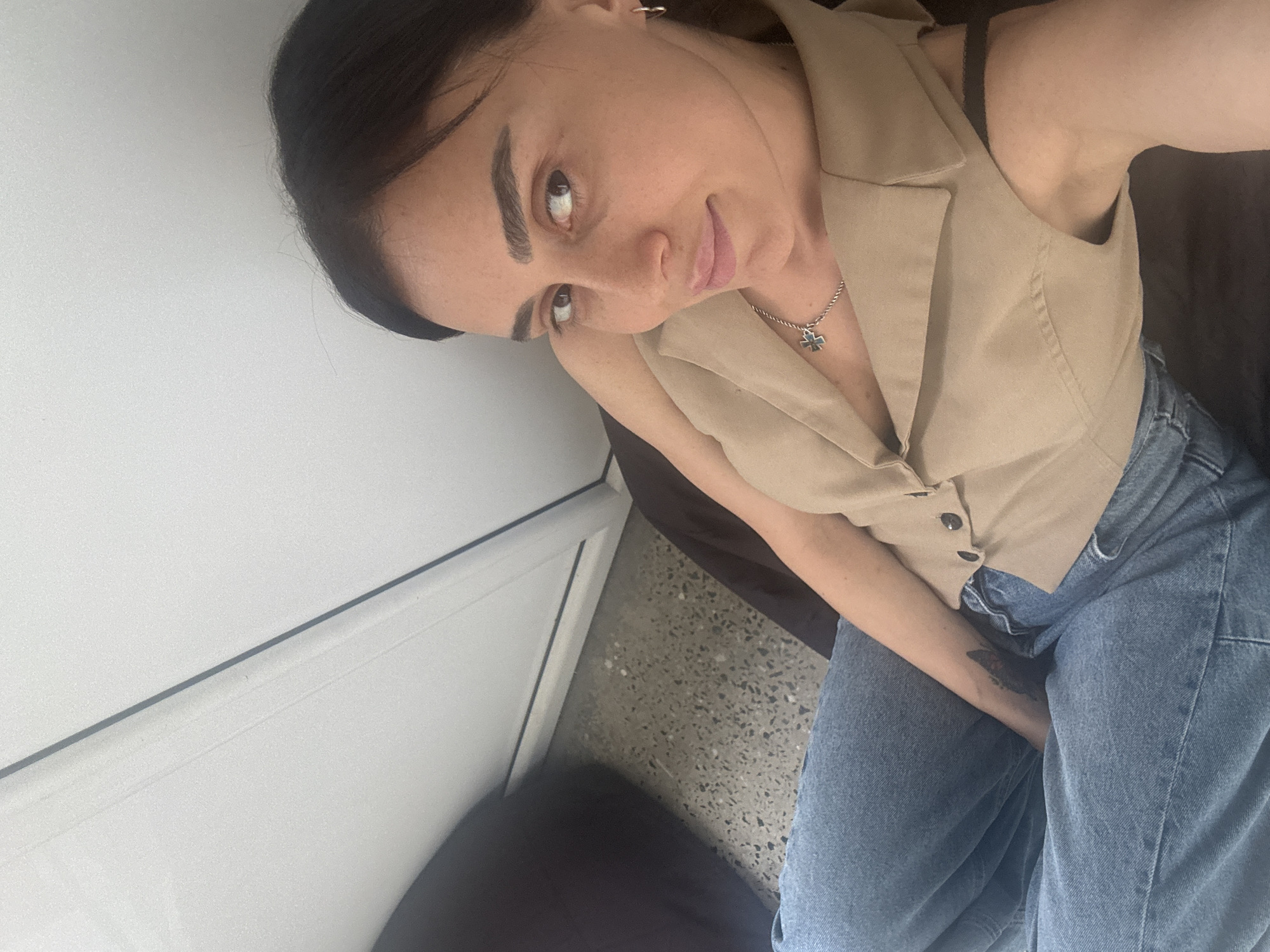 A medium shot captured from a high angle shows a woman sitting on a dark surface, possibly a chair or sofa, with her legs crossed in light blue denim jeans. She is wearing a beige vest over a dark top, and a silver necklace with a green cross pendant. Her right arm is bent and resting on her lap, revealing a butterfly tattoo on her forearm. To her left, a black object is partially visible, likely an armrest or cushion. The background consists of a wall with vertical lines and a textured floor, which is common in many modern buildings and public spaces in Tbilisi, Georgia. The lighting is soft and natural, suggesting an indoor setting during the daytime.