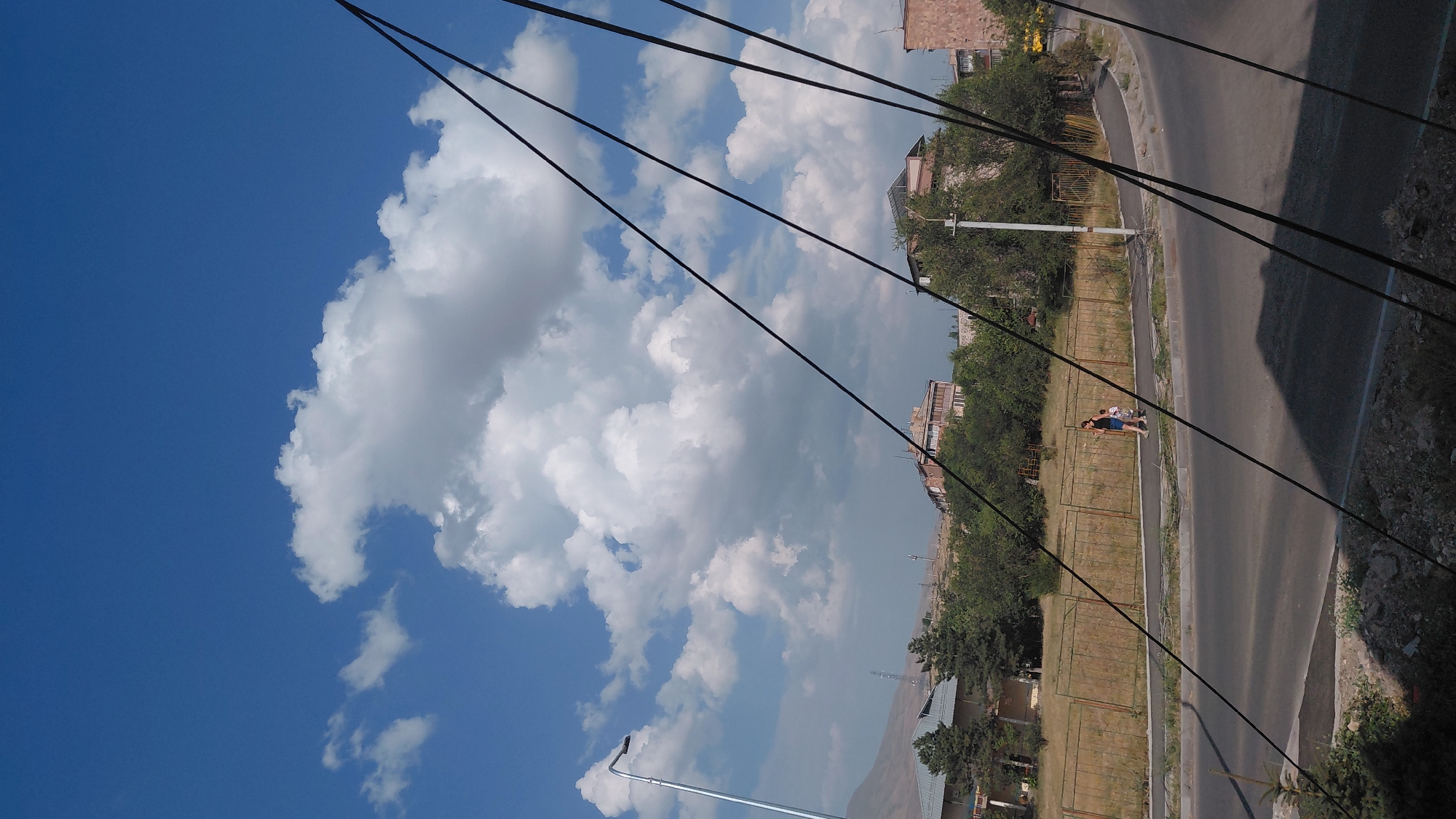 The image depicts a street scene in Abovyan, Armenia, under a bright blue sky with scattered white clouds. In the foreground, several dark, angled power lines dominate the frame, casting shadows on the asphalt road below. A sidewalk runs alongside the road, where a woman in a dark top and blue shorts and a child in a light-colored outfit are walking away from the camera. Behind the sidewalk is a grassy area with a chain-link fence. In the middle ground, trees and several multi-story buildings are visible, some appearing residential. A lamppost stands near the sidewalk. In the background, a mountainous landscape can be seen in the distance. The overall impression is an ordinary day in a residential area of the city.