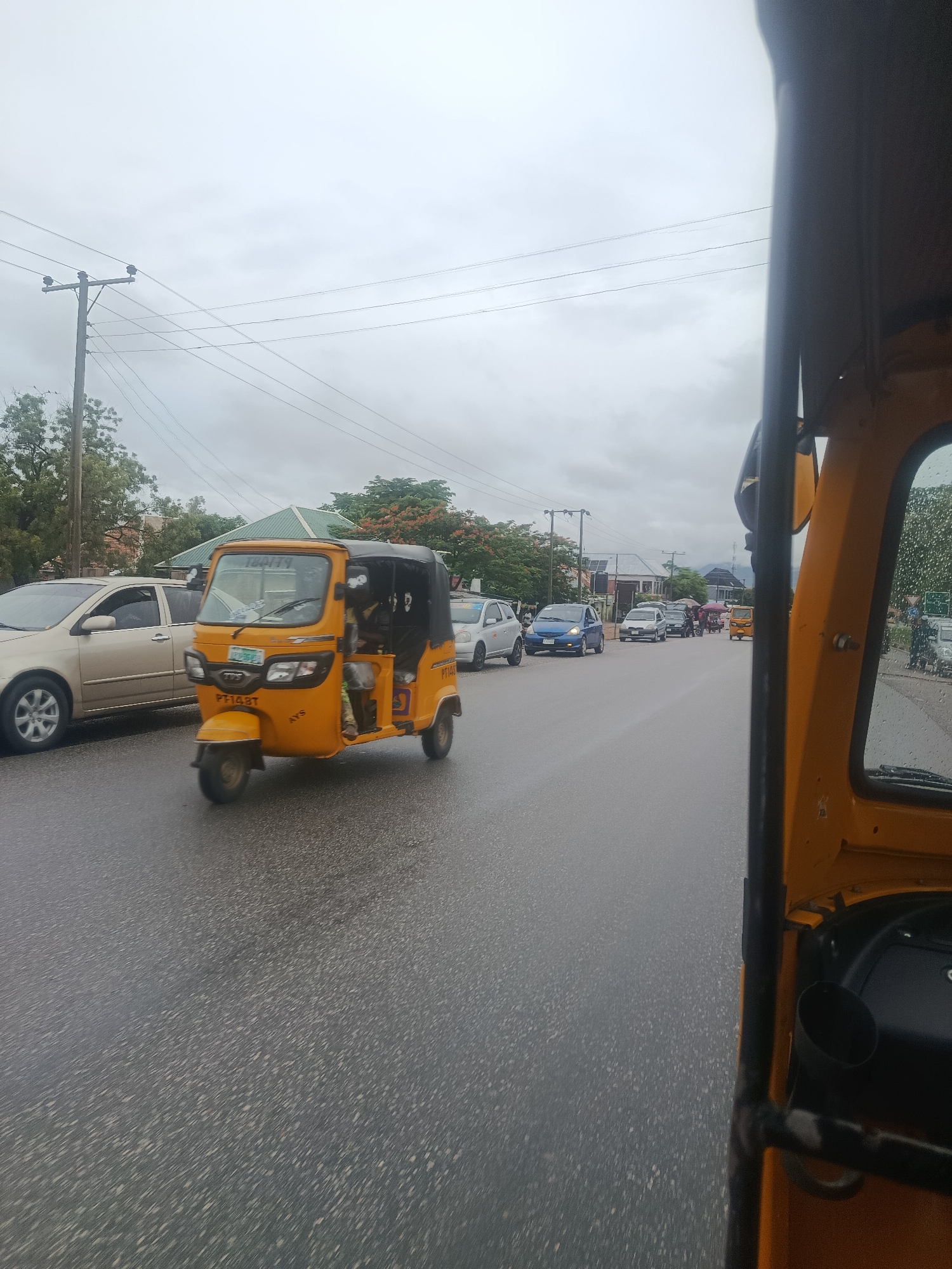 The image depicts a street scene in Jalingo, Nigeria, on a cloudy day. The foreground is dominated by the interior of a yellow auto-rickshaw, with a black frame and a portion of the yellow body visible. The road ahead is wet, suggesting recent rain, and several vehicles are in motion. A yellow auto-rickshaw, similar to the one from which the photo is taken, is in the mid-ground, with a driver and a passenger visible inside. Following this are several cars, including a silver sedan, a blue hatchback, a darker sedan, and other vehicles further down the road. Trees line the sides of the street, with some buildings visible in the background. Utility poles with electrical wires are also present. The overall impression is that of a moderately busy street with ongoing traffic.