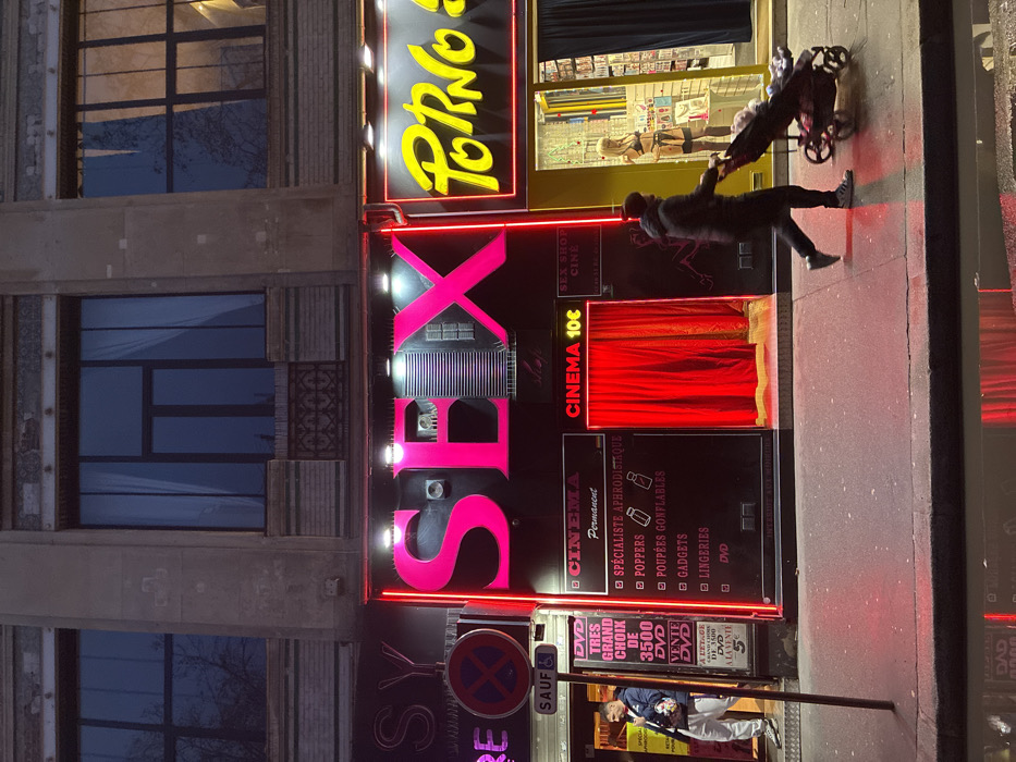 The image shows the exterior of a sex shop in an urban area. Neon signs spell out "SEX," "PORNO," and "CINEMA 10€." The shop advertises various products, including DVDs, lingerie, gadgets, and aphrodisiacs. A red curtain is visible behind a window. A person pushes a stroller in front of the shop. The building's facade is adjacent to the sex shop. A pedestrian is visible near a post with prohibition symbols.
