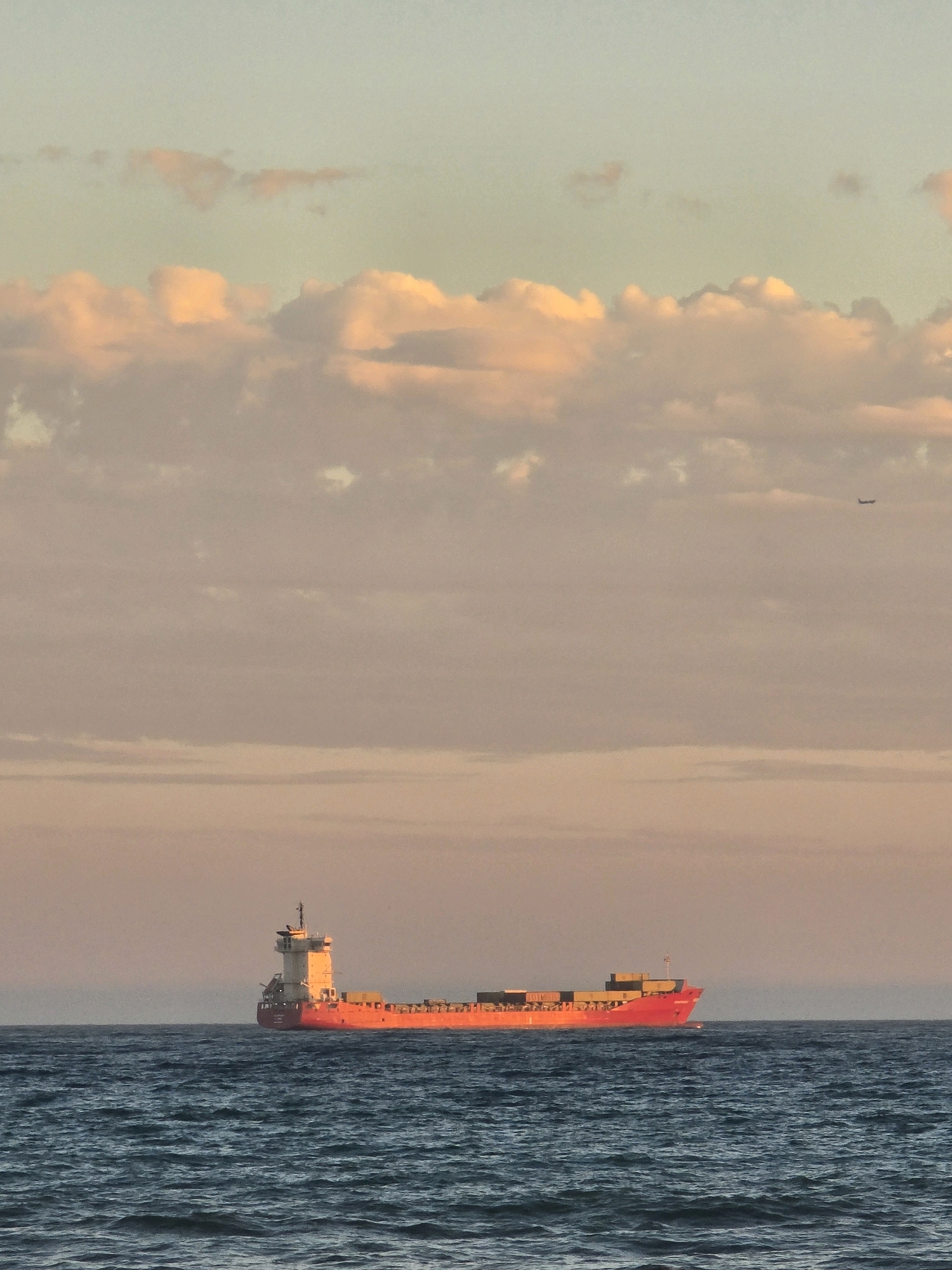 A cargo ship sails on the sea under a dusky sky. The ship is bright orange and filled with containers. The sky is a soft gradient of blue and pink with scattered clouds. A small airplane is visible in the upper right corner of the image. The sea is a deep blue with moderate waves.