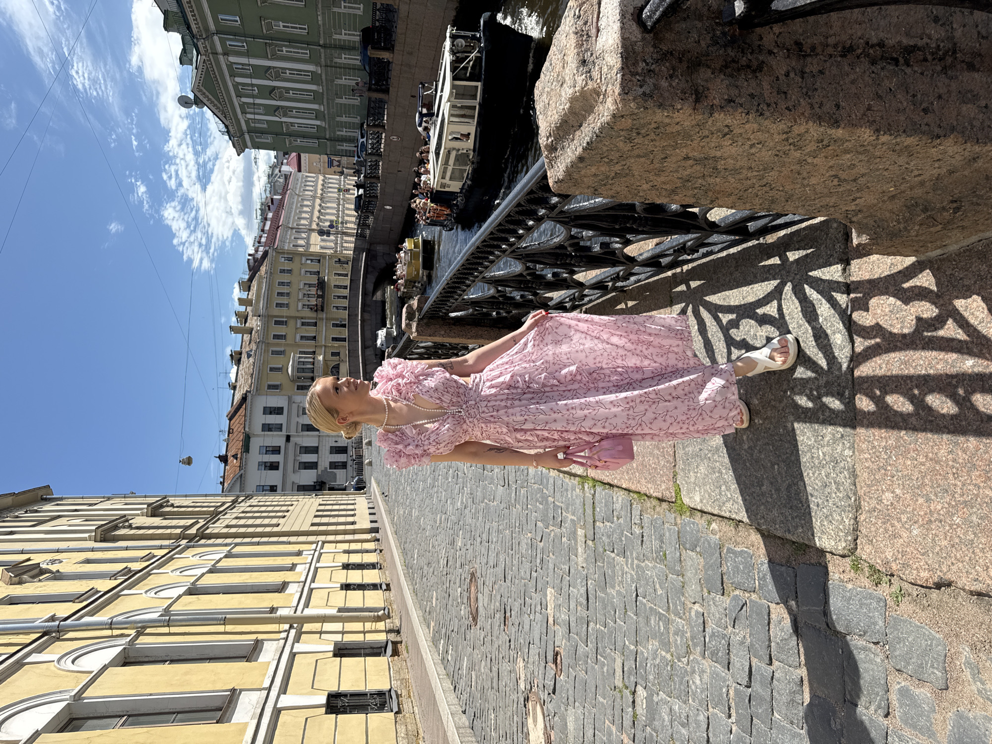 A young Caucasian woman with blonde hair pulled back in a bun, wearing a pink floral print maxi dress with ruffled sleeves and a plunging neckline, and white platform sandals, walks along a cobblestone sidewalk next to a canal in Saint Petersburg, Russia. She is carrying a small pink handbag. The canal is lined with a black metal railing. Two tour boats are on the water, one with passengers, and a bridge is visible in the background. The buildings on either side of the canal are old, European-style architecture, with one building on the left being yellow and the building on the right being green. The sky is blue with scattered white clouds, and the sun casts shadows on the sidewalk, indicating it is daytime.