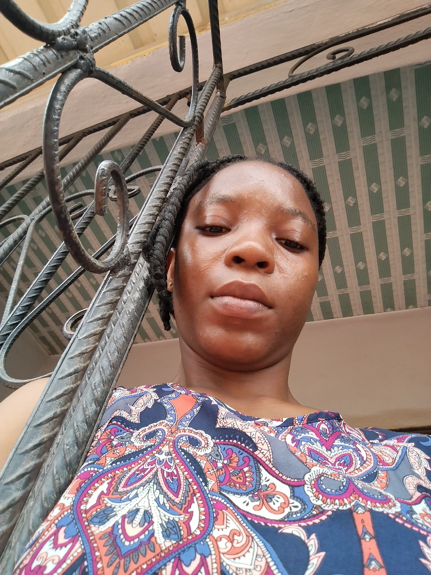 A young woman, likely of Nigerian descent, is pictured outdoors in Isiehor, Nigeria. She is leaning against a metal railing, possibly part of a gate or fence. The woman has dark skin and her hair is braided. She is wearing a colorful blouse with an intricate paisley pattern in shades of blue, pink, orange, and white. Behind her, a patterned ceiling with green and white stripes is visible, along with a section of a beige wall. The perspective of the image is a low-angle shot, looking up at the woman. The lighting suggests it is daytime.