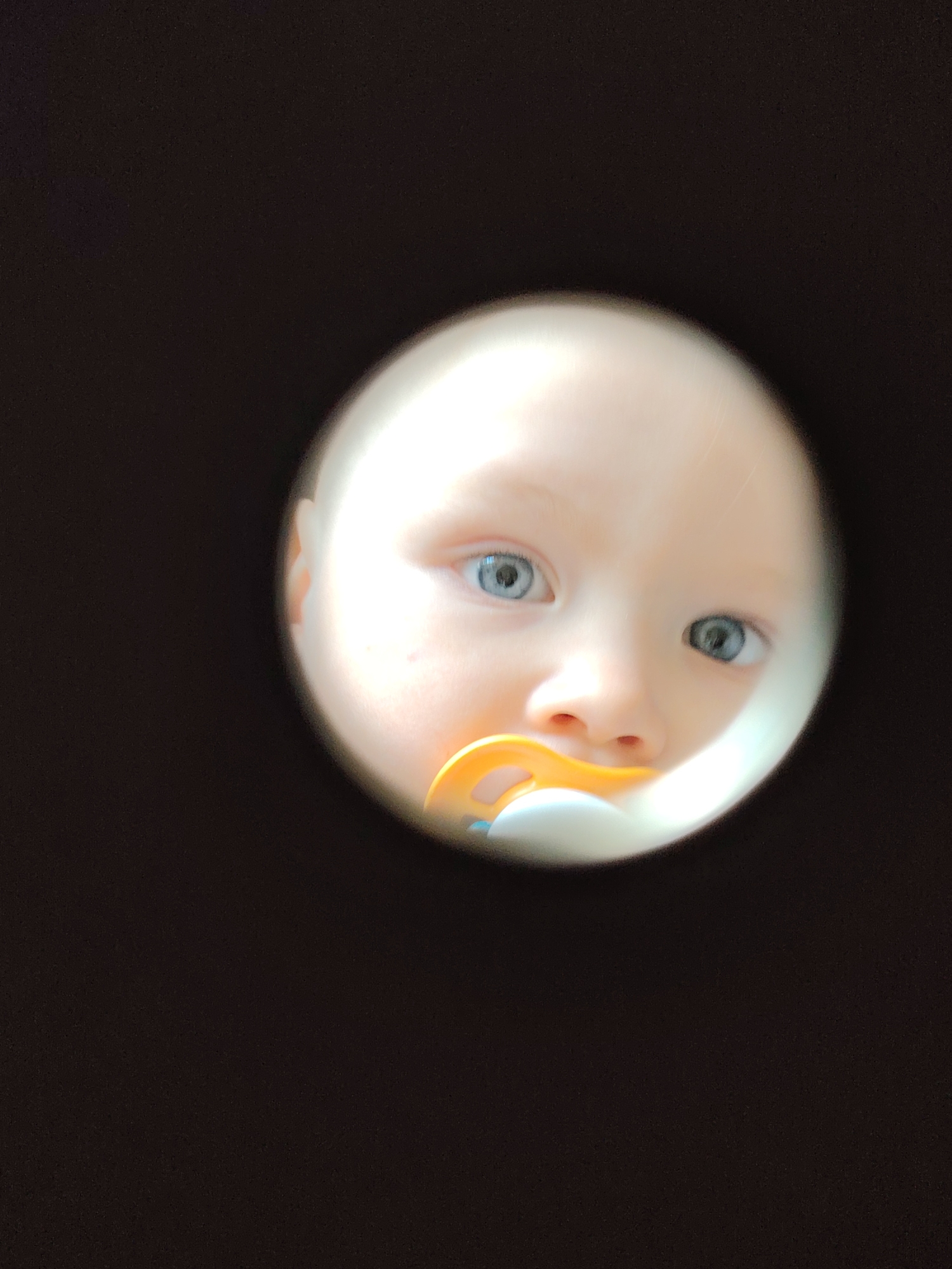 A baby with blue eyes and a pacifier is visible through a circular opening in a dark surface. The location is unknown.