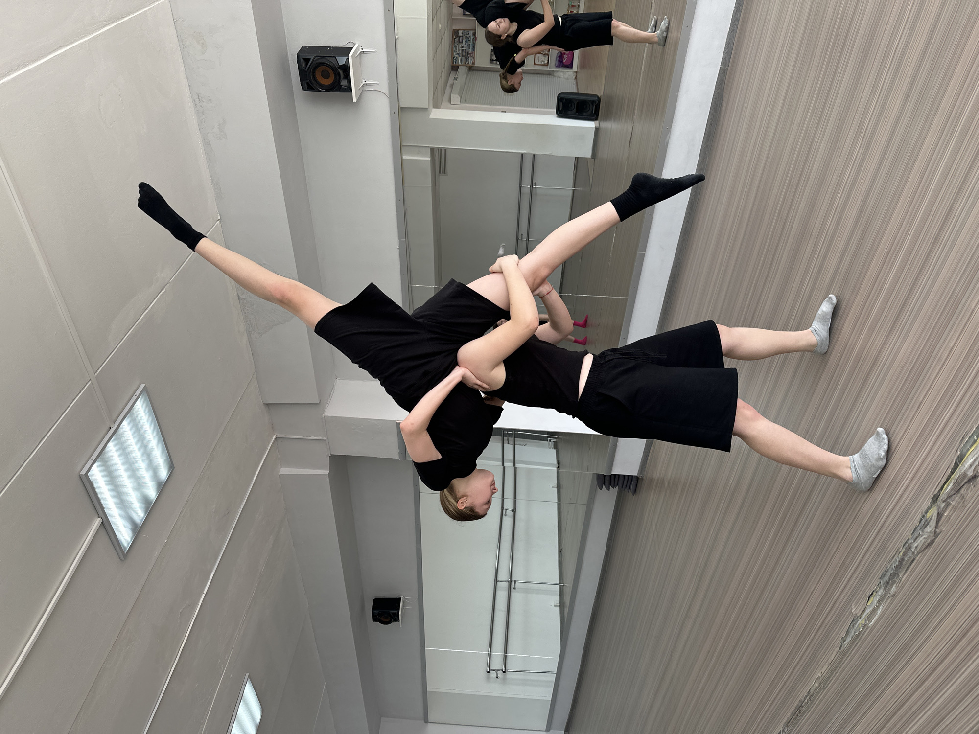 Two individuals in black athletic wear are engaged in a dance lift in a studio or dance classroom. One person is supporting the other, who is extended horizontally with one leg pointing upward. The room features a mirror on one wall, reflecting the scene, and a light fixture on the ceiling. The floor has a wood-like finish. A speaker is mounted on the wall. The location and city are not identifiable from the image.